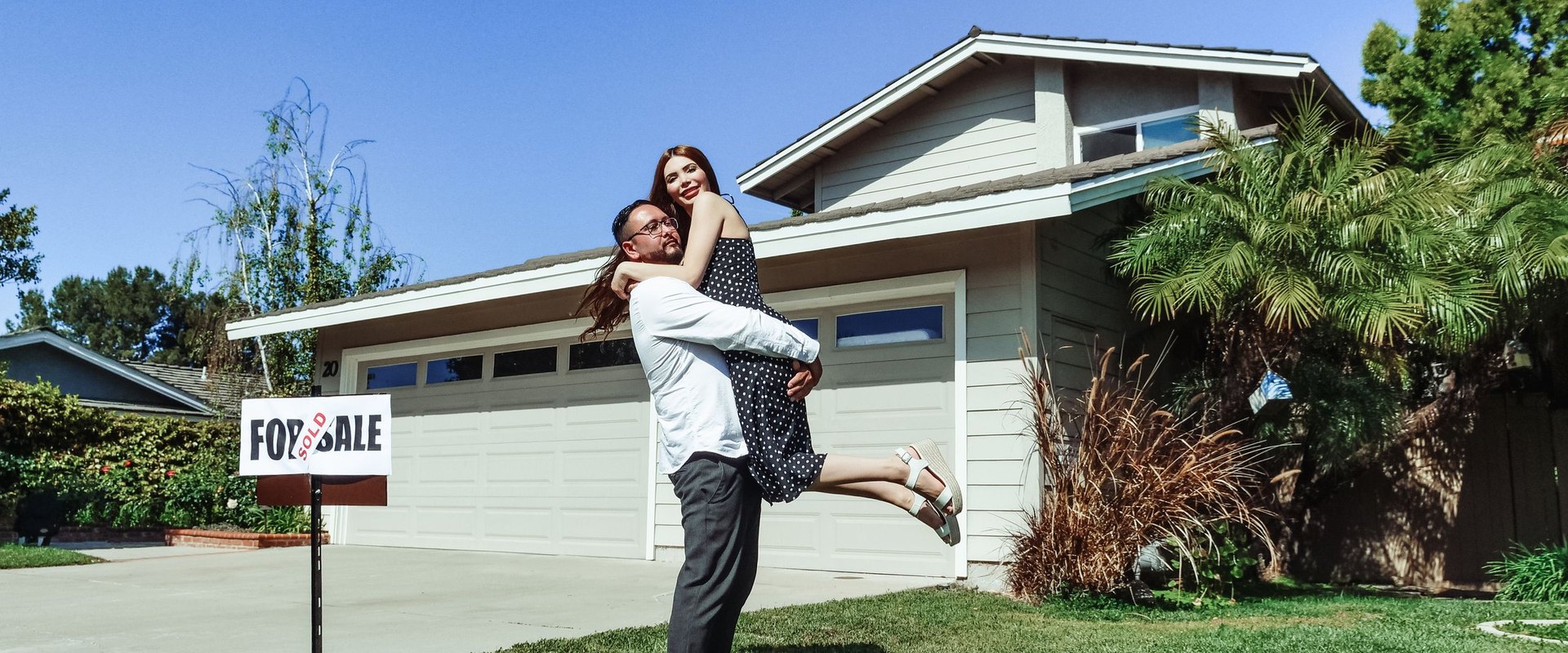 A couple celebrates in front of a single-story home with a For Sale sign and a Sold rider in the yard.