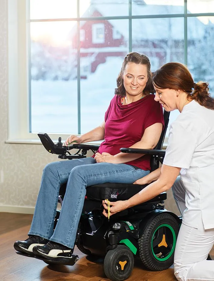 Woman in power wheelchair receiving personalized fitting assistance from a caregiver, emphasizing mobility solutions and comfort in a home setting.