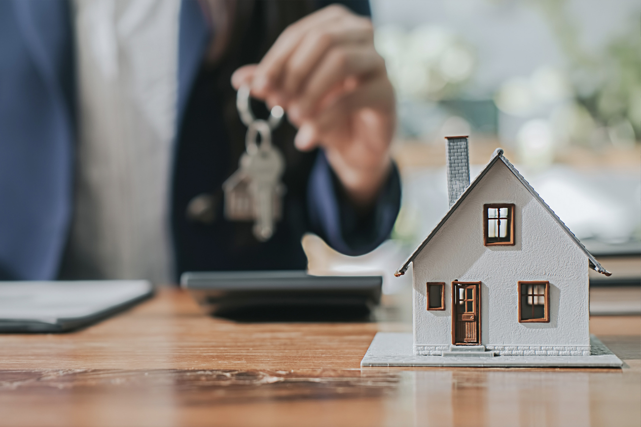 Person holding house keys above a model home on a wooden desk, symbolizing real estate transactions and property ownership in Wharton County, Texas.