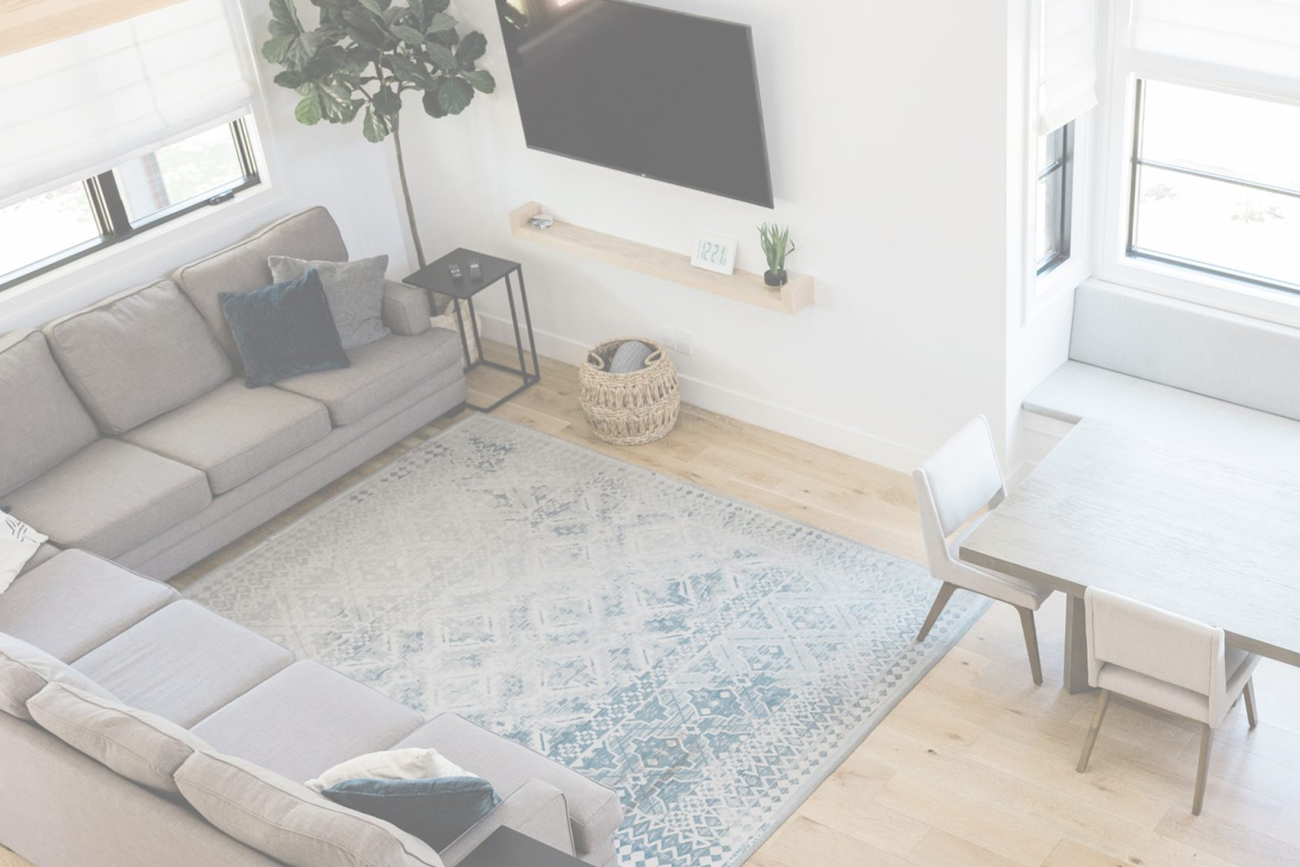 Modern living room featuring a gray sectional sofa, decorative pillows, a textured area rug with blue accents, a wooden coffee table, and a small plant next to a wall-mounted TV, highlighting a bright and airy space ideal for relaxing or entertaining.