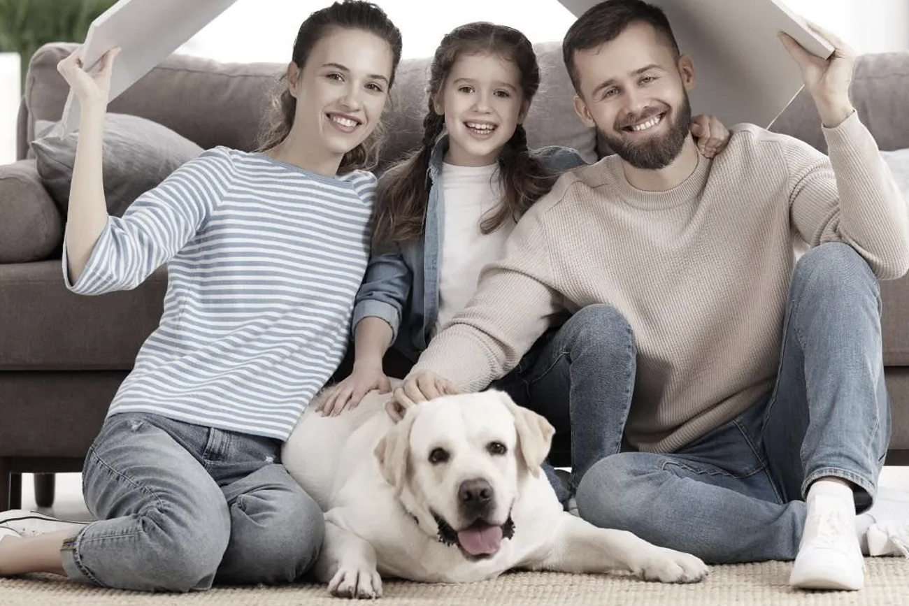 Family of three with a yellow Labrador dog, sitting on a rug in a cozy living room, smiling and enjoying time together.