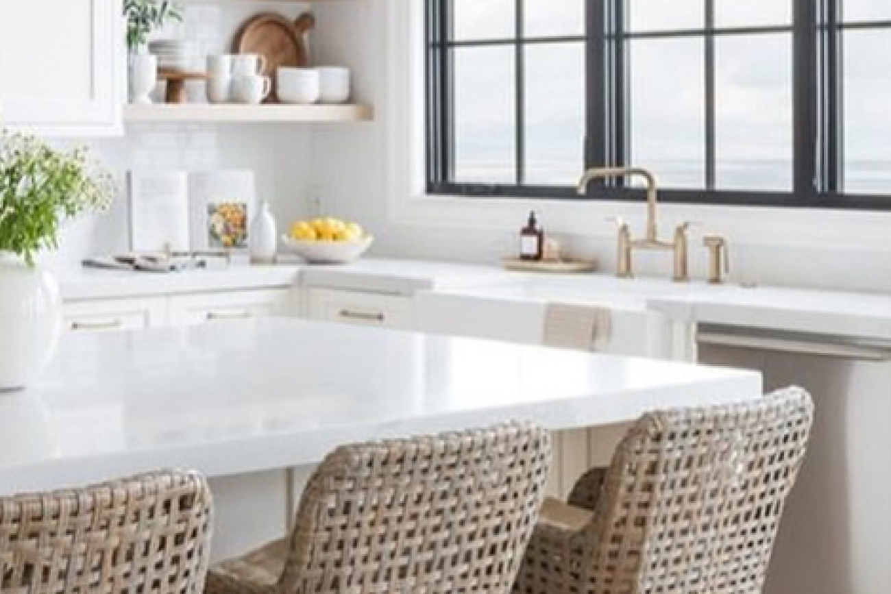 Modern kitchen with white cabinetry, marble countertop, and woven chairs, featuring a bowl of lemons, decorative dishware, and large windows providing natural light.