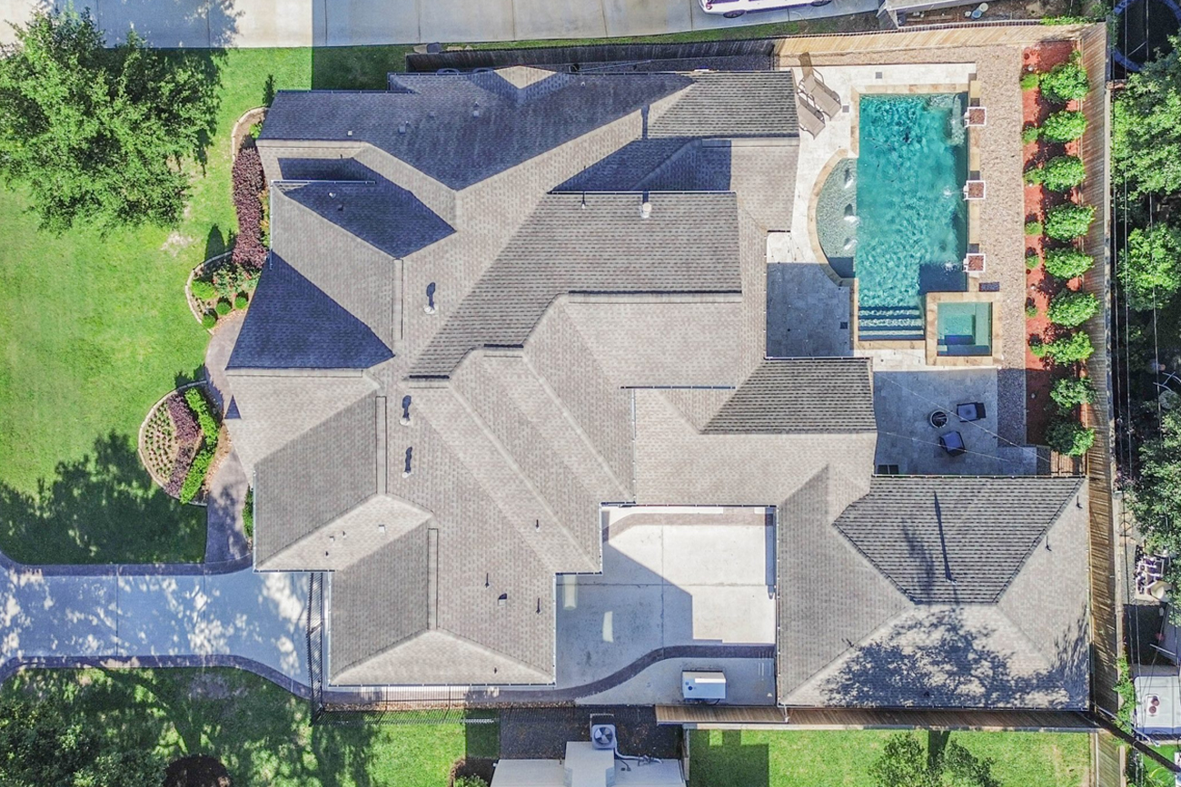 Aerial view of a spacious residential property in Austin County, Texas, featuring a modern roof design, landscaped yard, and a swimming pool surrounded by patio areas.