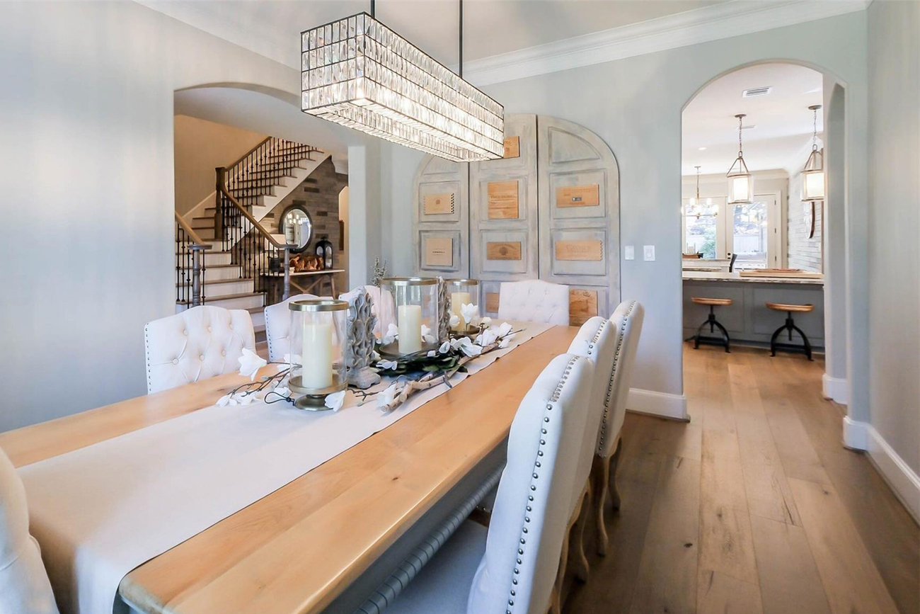 Elegant dining room featuring a long wooden table with white tufted chairs, decorative candles, and a stylish chandelier, leading to an open kitchen area with bar seating and modern decor.