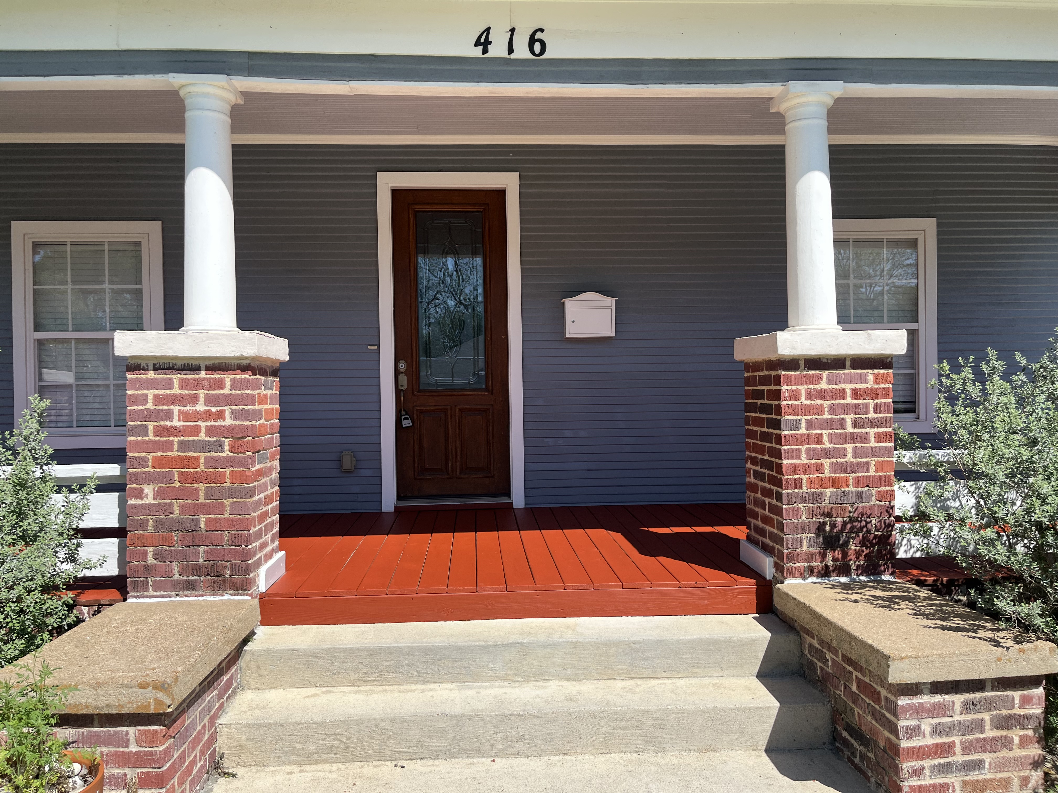 Front porch with freshly painted red/terra cotta deck boards, gray siding, white trim, and classic white columns