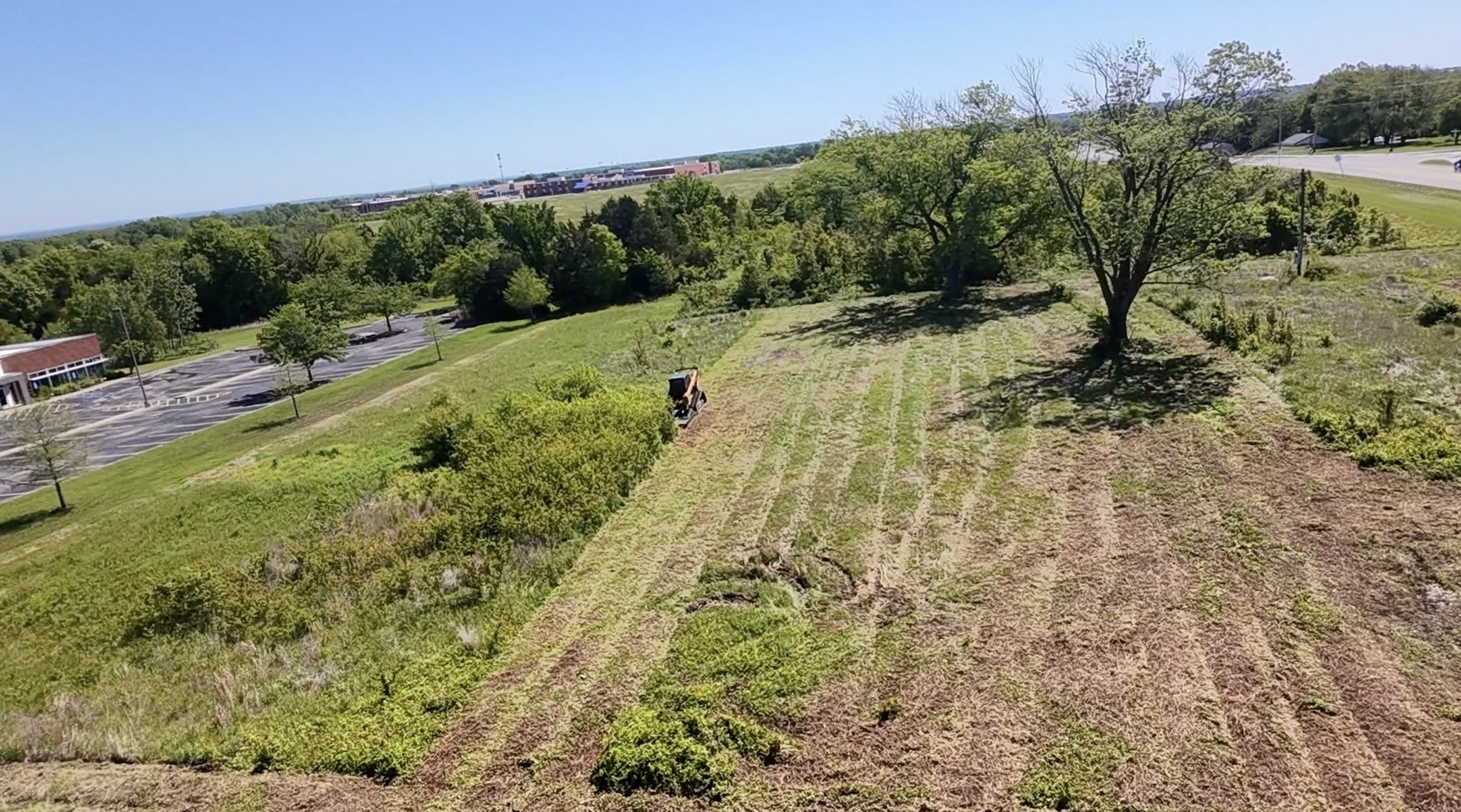 Aerial view of Kubota machine clearing overgrown field near commercial area
