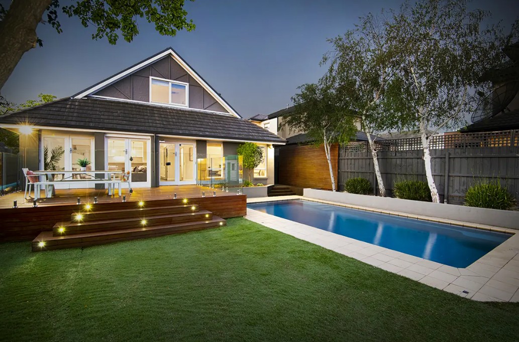 Twilight exterior of a charming home with pool, deck lighting and lush garden