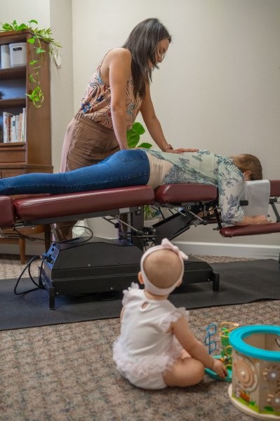 Dr. Liz evaluating spinal and pelvic mobility for a patient while a baby sits comfortably nearby at Mojica Chiropractic Care
