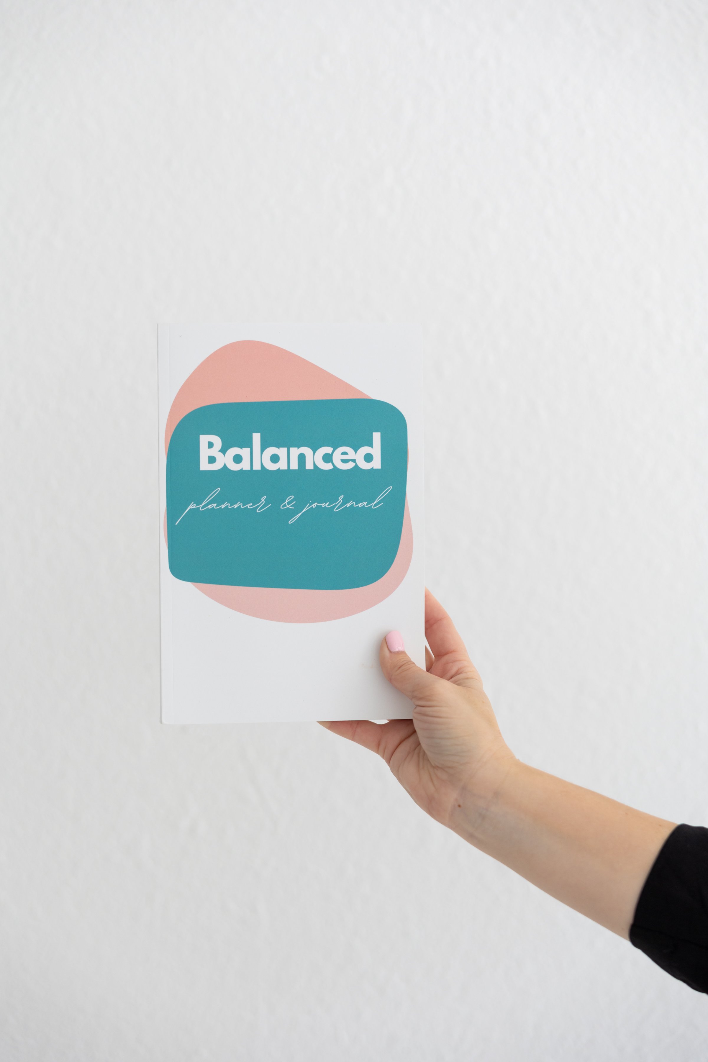 A hand holding a planner and journal titled 'Balanced' in front of a plain white wall.