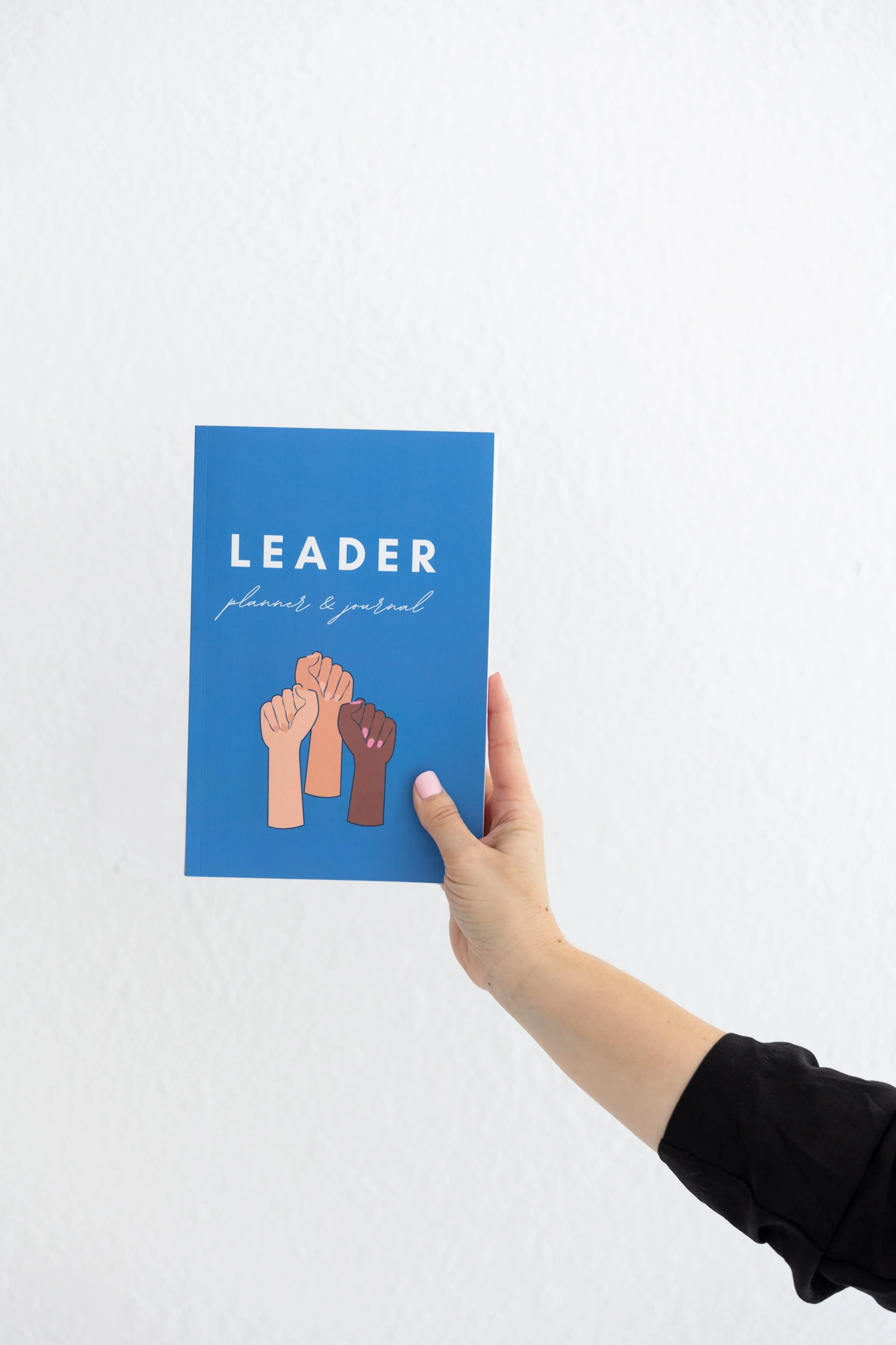 A hand holding a blue Leader Planner & Journal with illustrated raised fists on the cover.