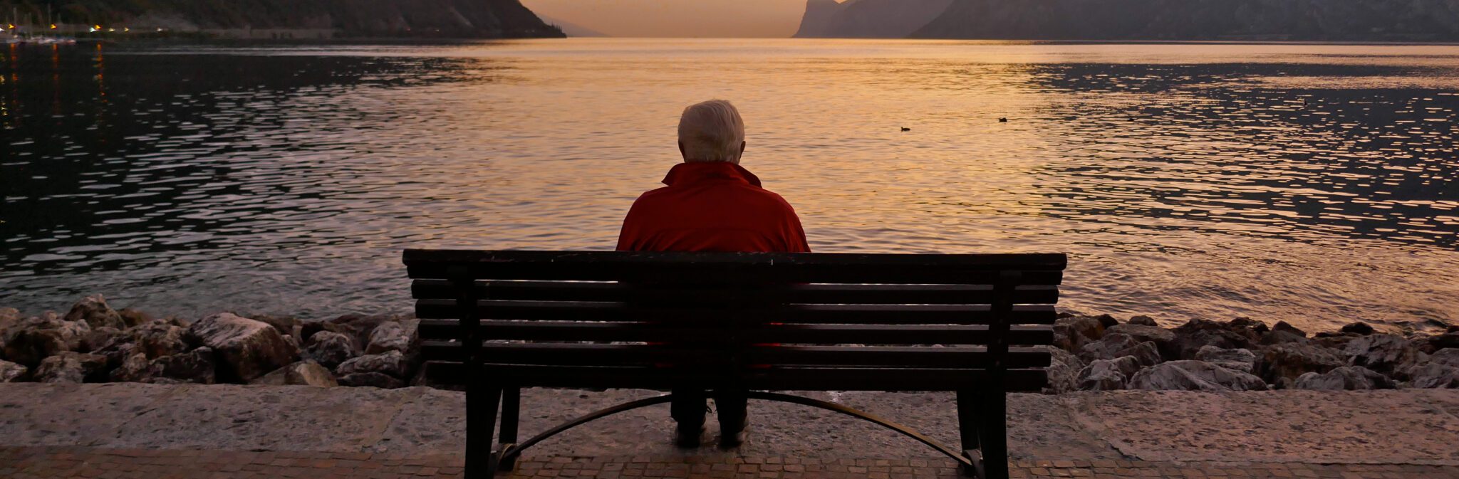 Older person sitting on a bench looking at the sunset over water