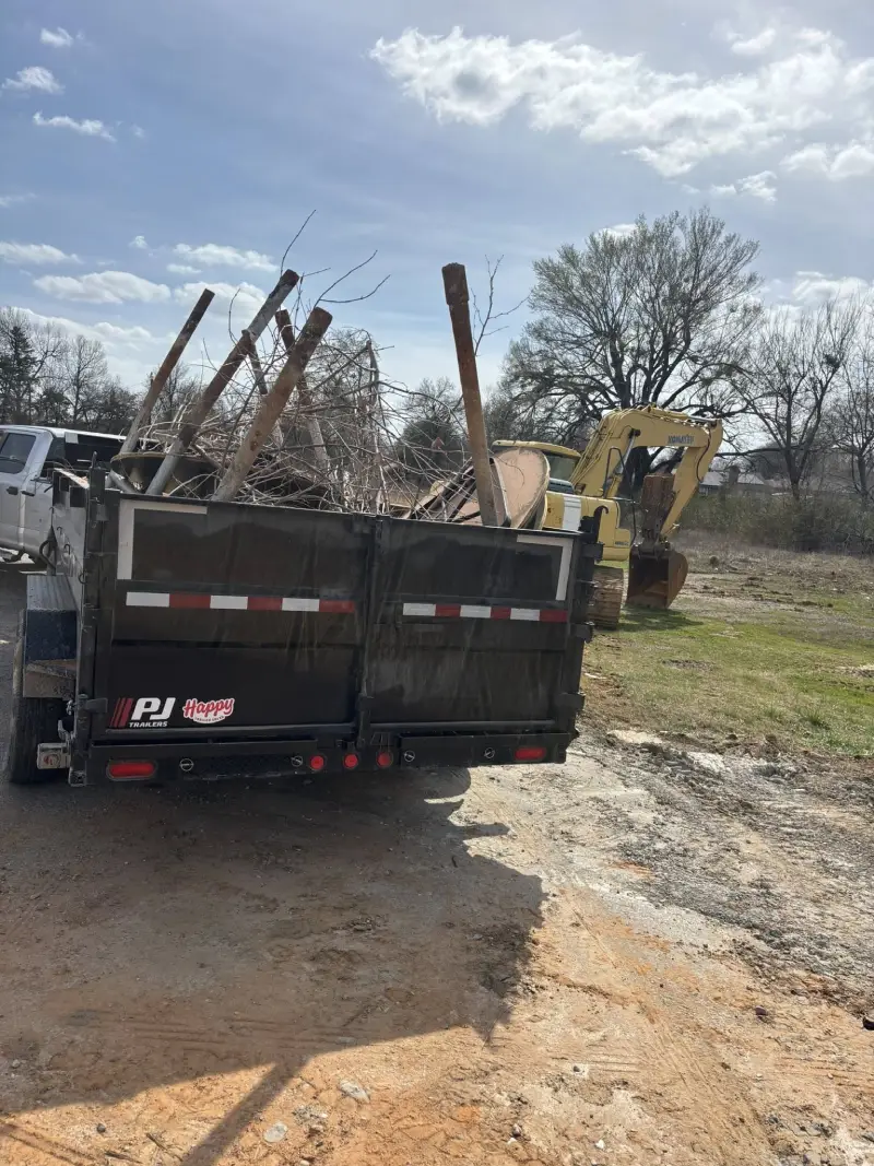 Dump trailer loaded with tree branches and an excavator in the background