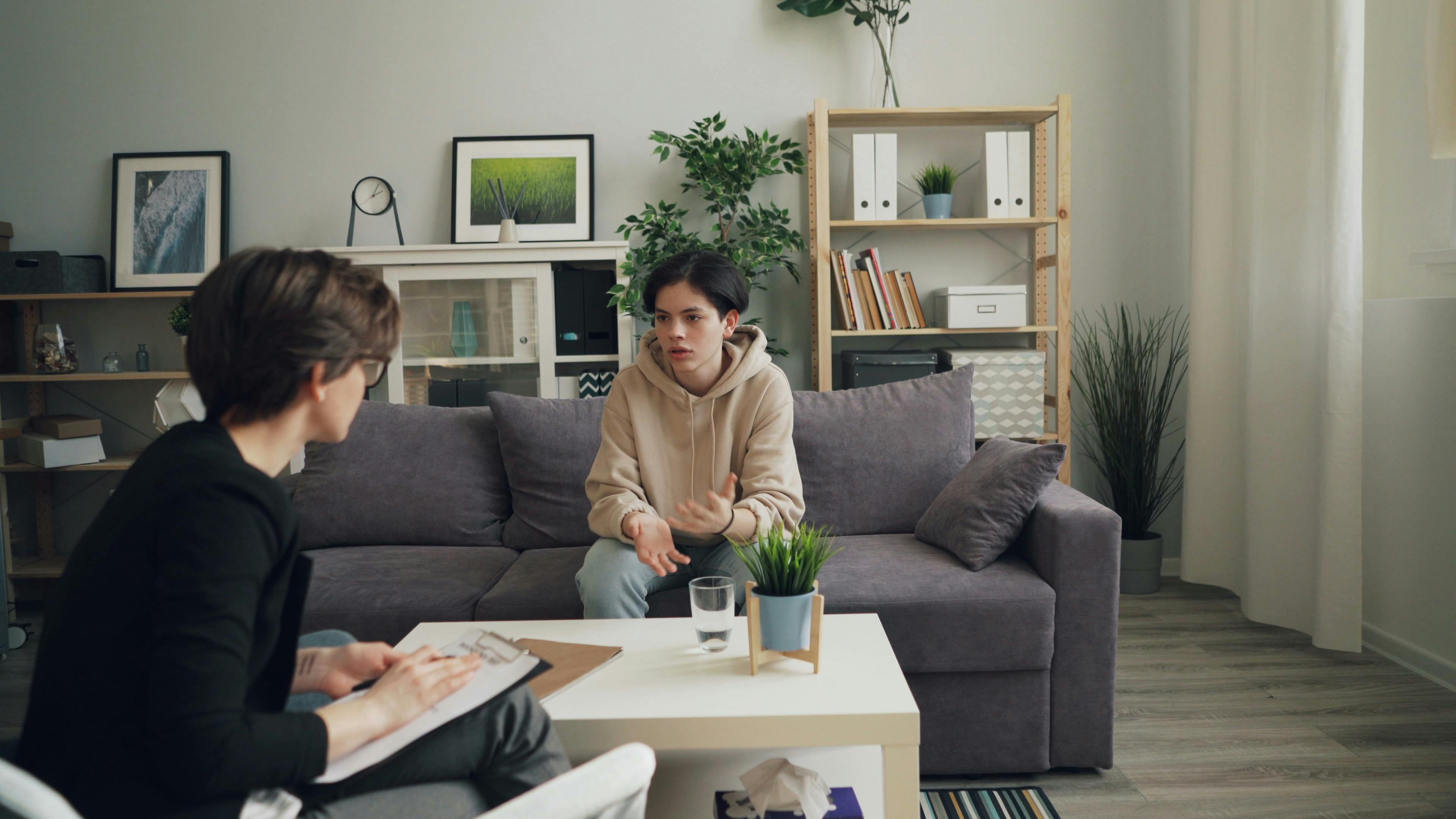 Individual counselling session in a cozy therapy room, featuring a young person in a beige hoodie discussing with a counsellor, with a notepad and glass of water on a coffee table.