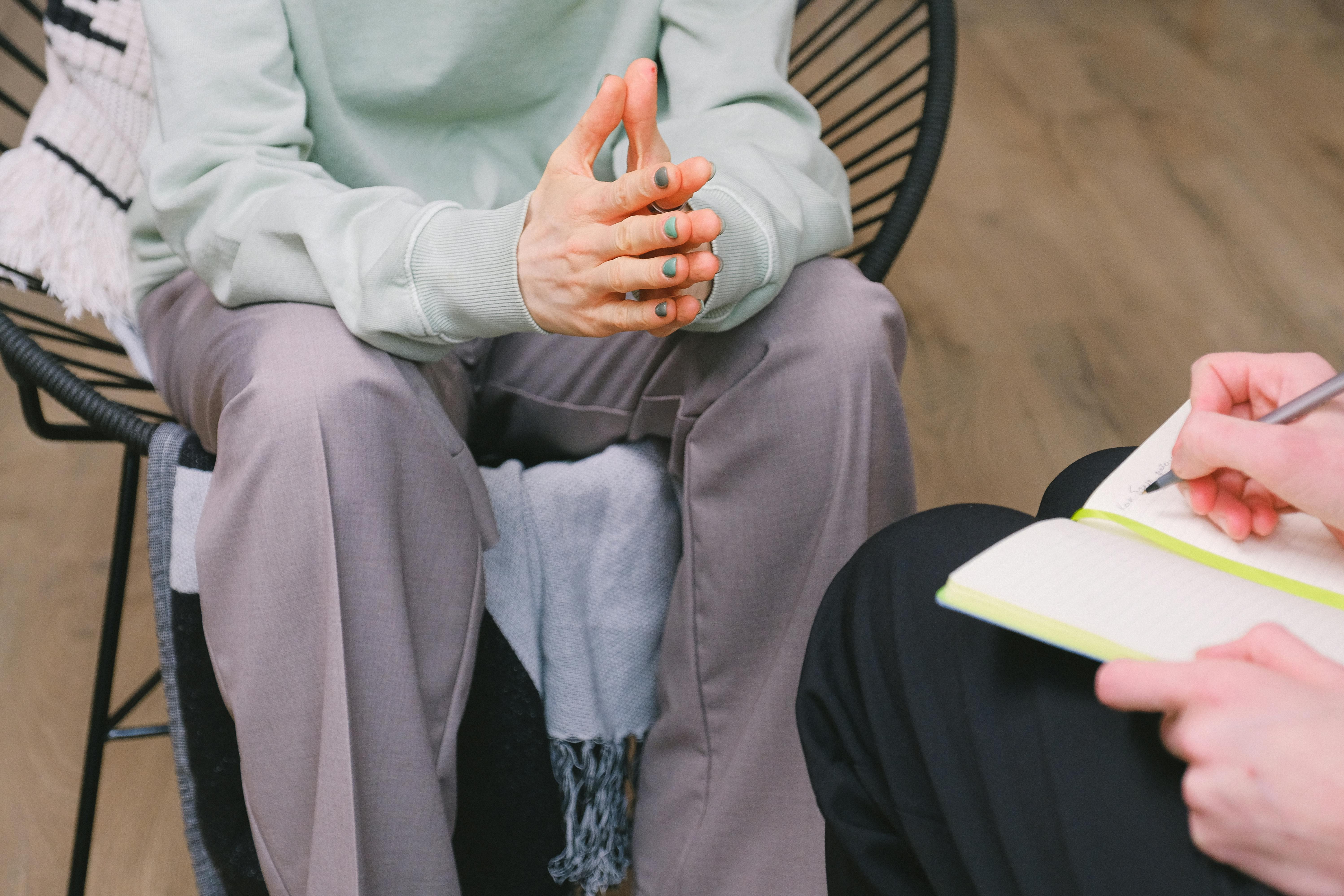 Person sitting in a chair with hands clasped, engaged in a counseling session, while another person takes notes in a notebook, reflecting the therapeutic environment of EMDR therapy for trauma and PTSD.