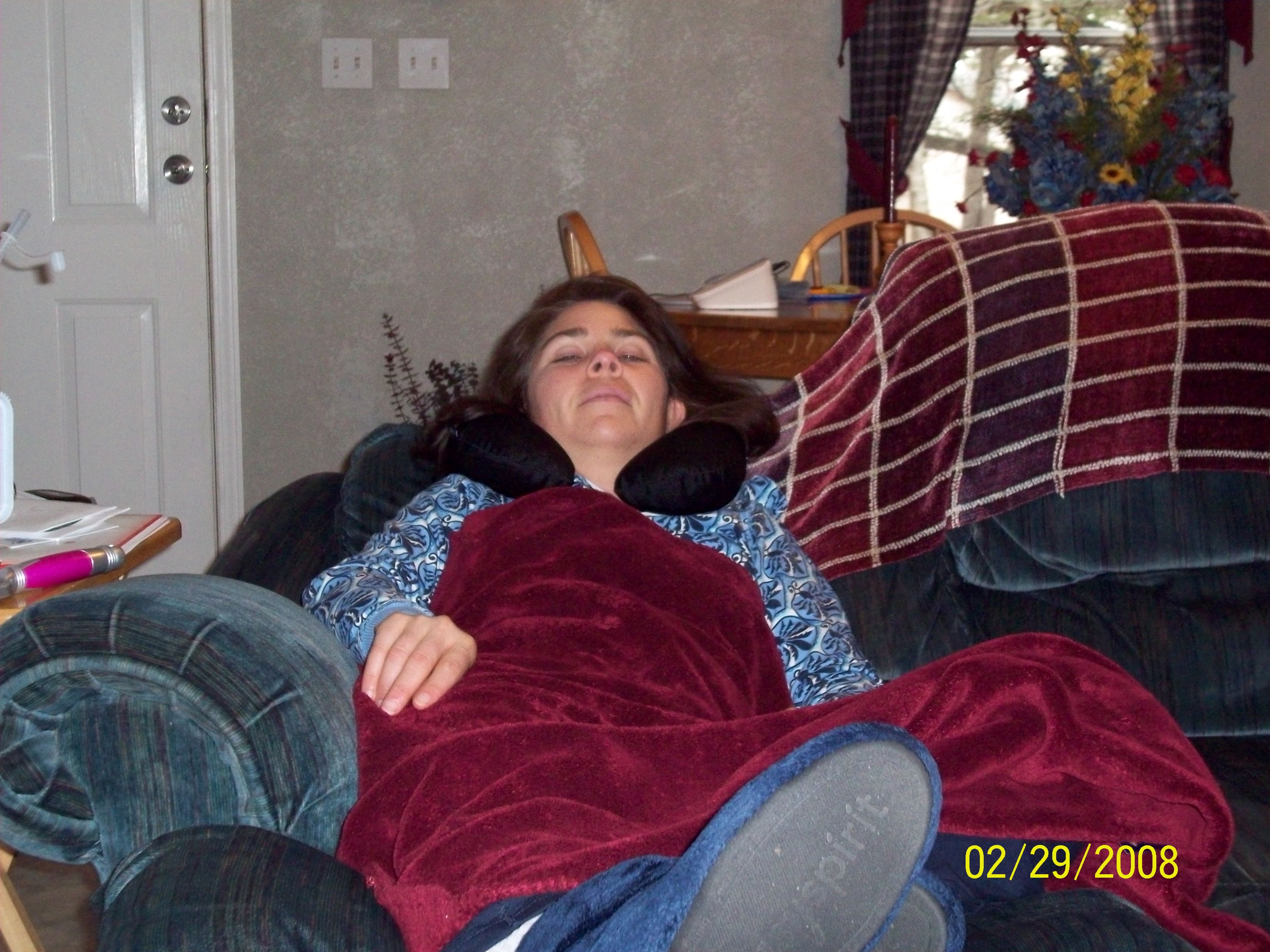 Woman resting on a couch, covered with a blanket, wearing a neck pillow, illustrating fatigue and chronic illness struggles.