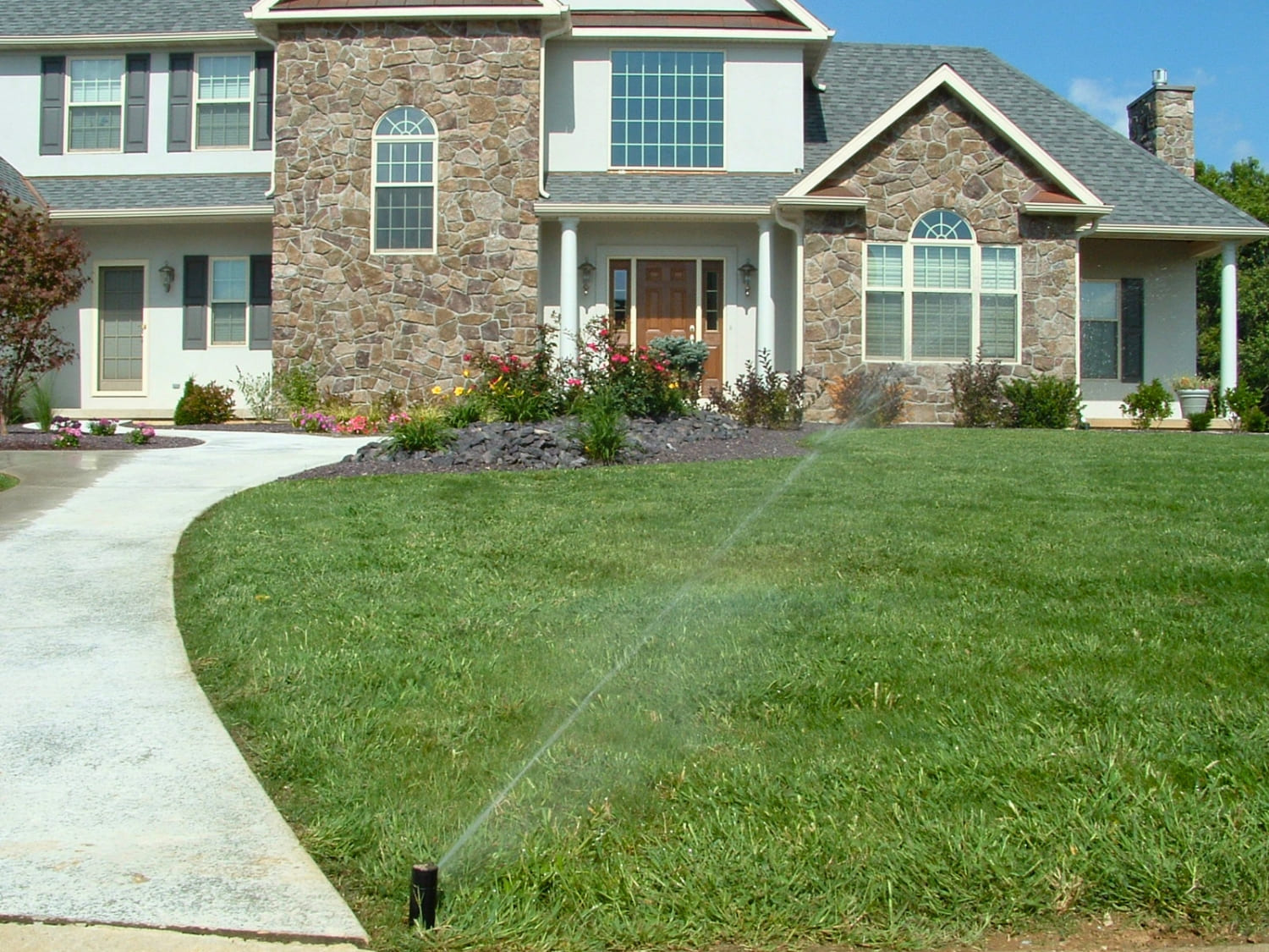 Residential lawn sprinkler system watering grass in front of a stone house