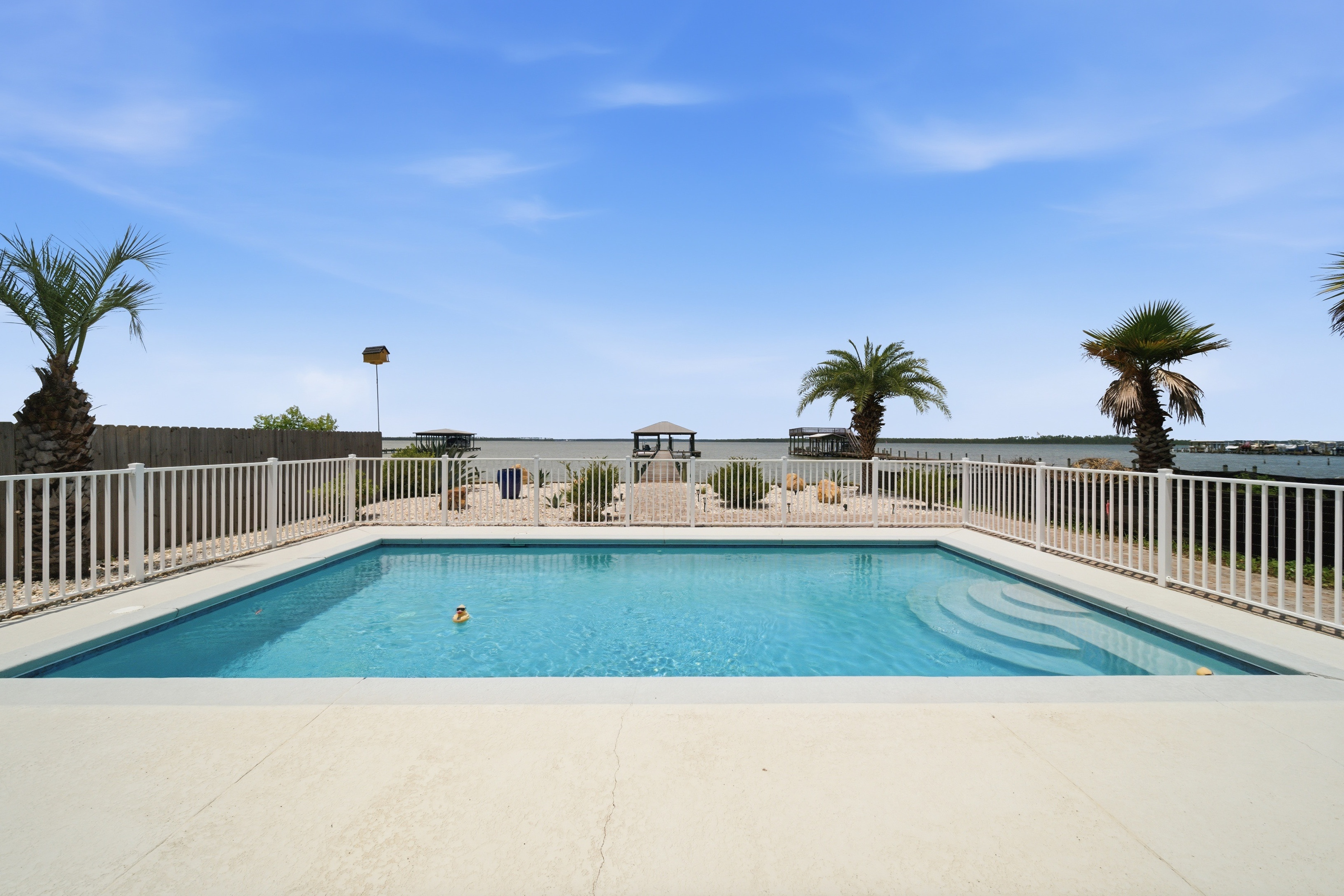 Private pool with water views, palm trees, and boat dock beyond the fence