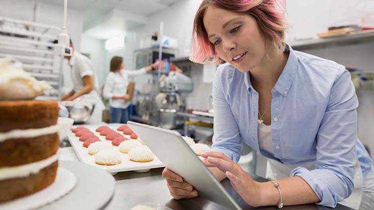 Small business owner reviewing payroll and workforce schedule on a tablet in a commercial bakery kitchen