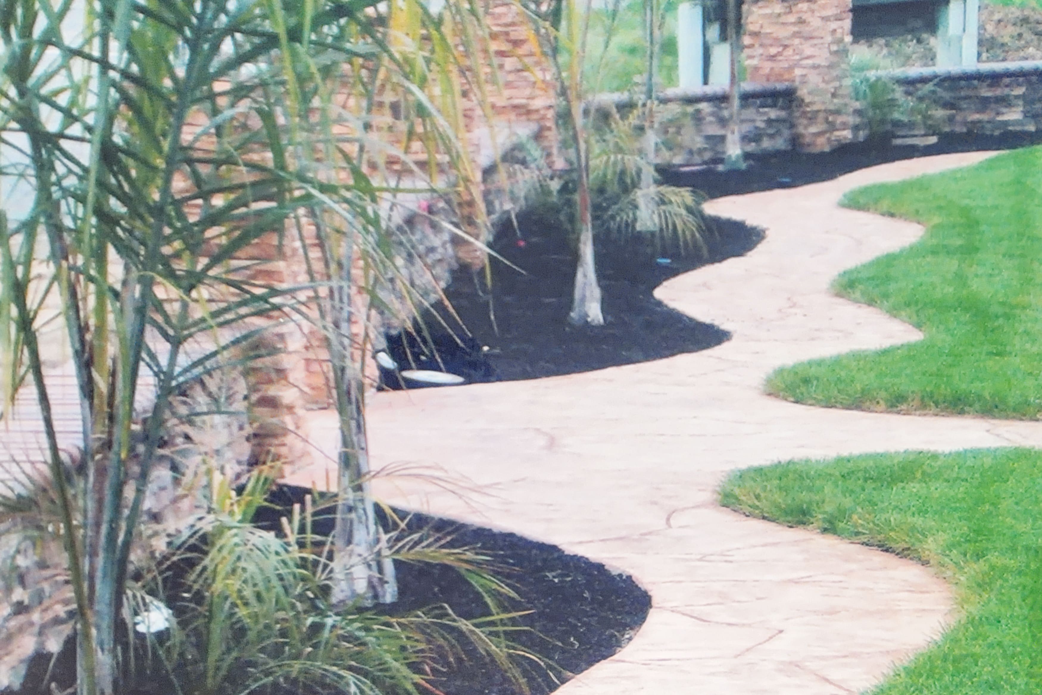 A winding stamped concrete pathway through a landscaped area with palm trees and a stone retaining wall