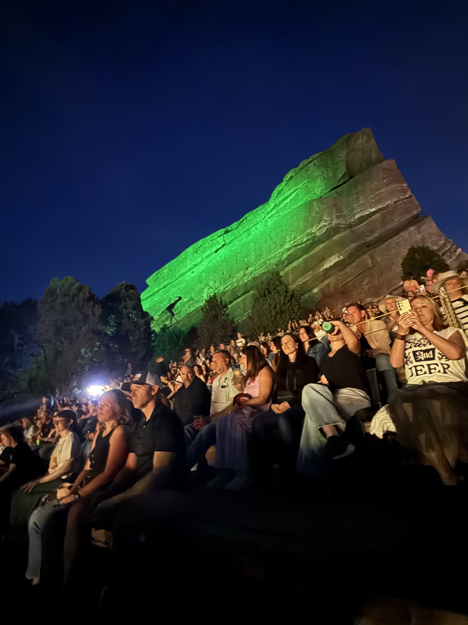 Audience seated beneath the massive rock face washed in vibrant green light