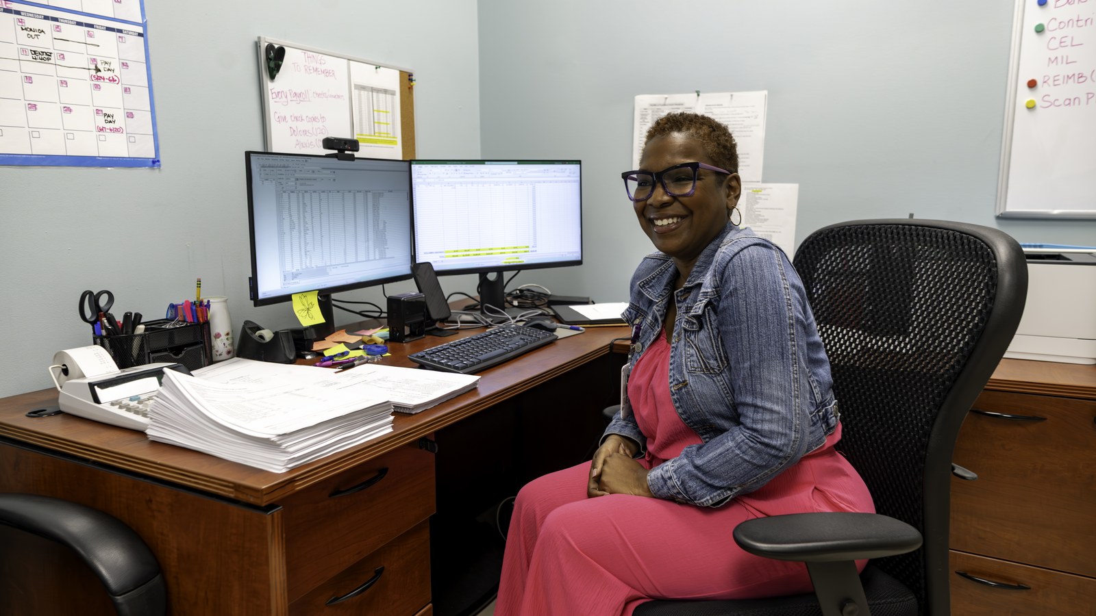 A staff member smiling at their desk in a non-profit office with dual monitors.