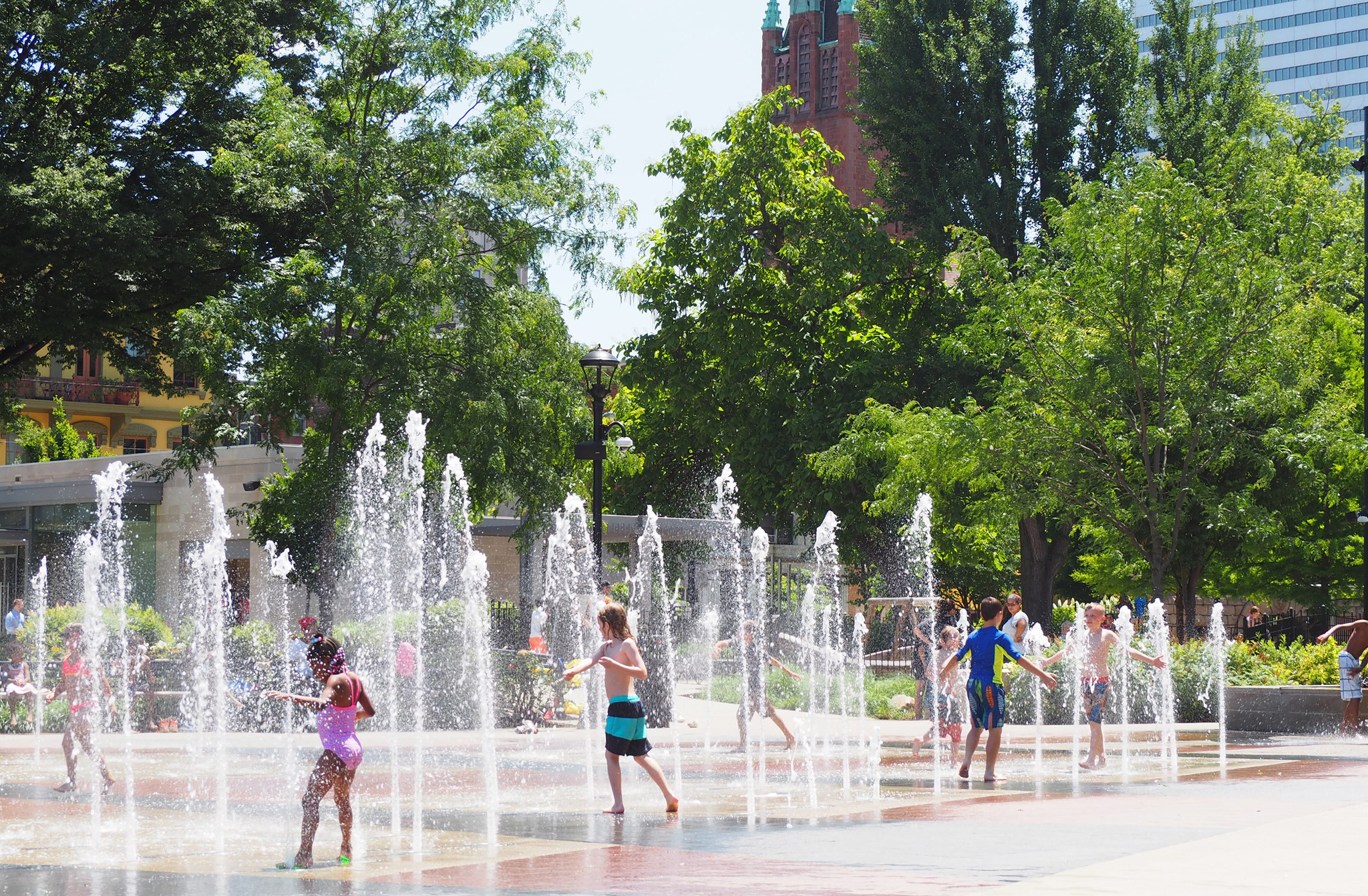 Families and children playing in a modern urban splash pad fountain in a lush city park on a sunny summer day