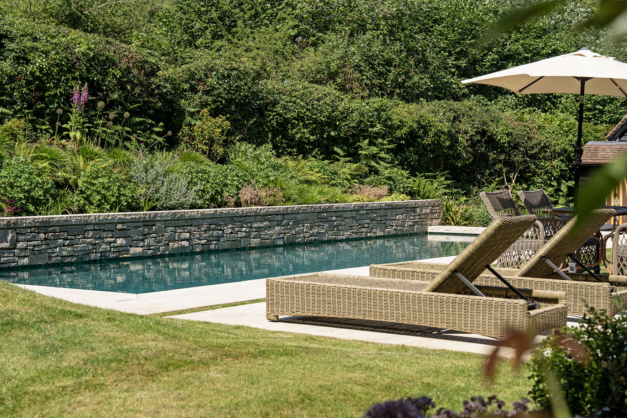 Luxury garden swimming pool with natural stone retaining wall and sun terrace by Poolcourt