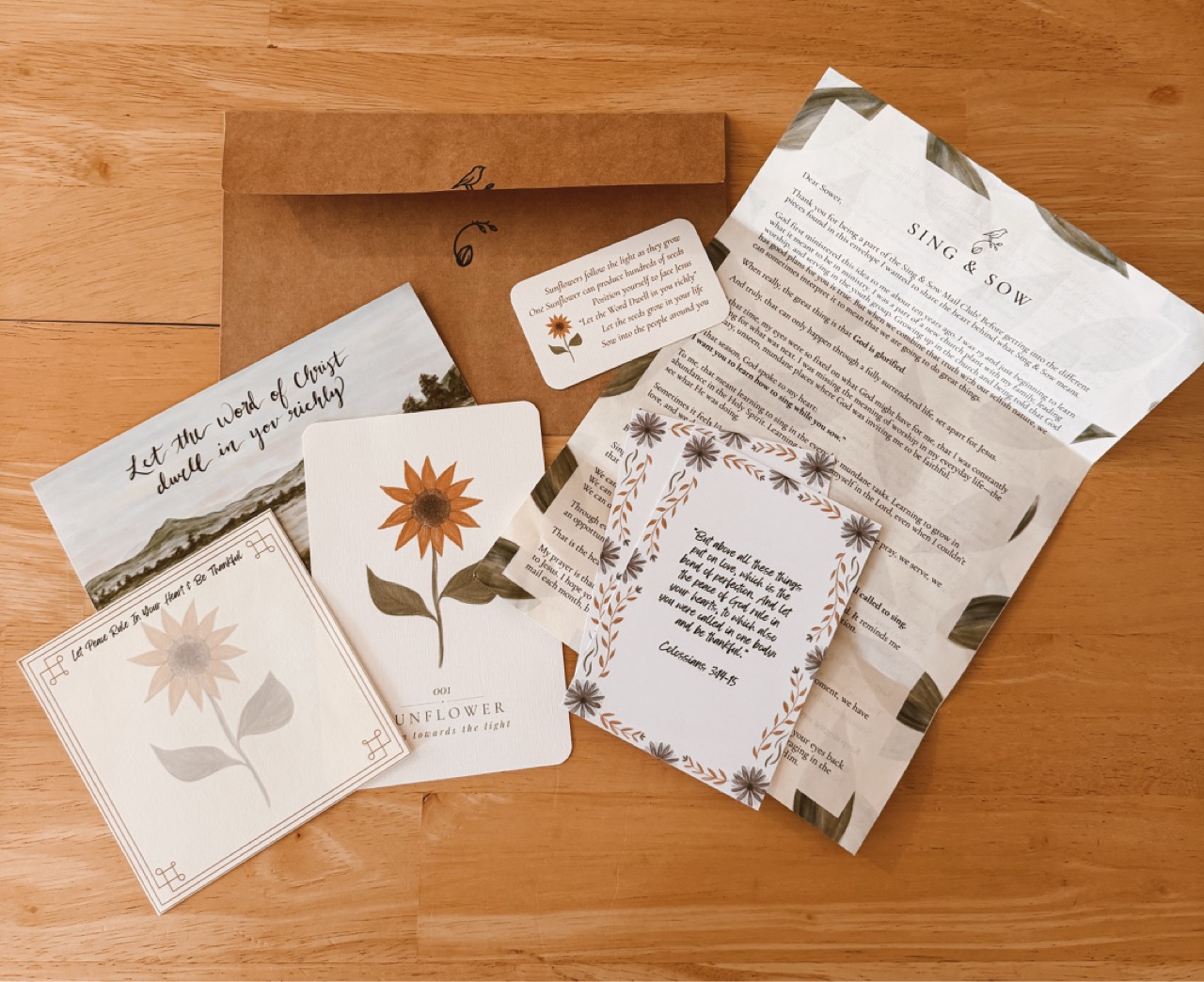 A past Mail Club package: a sunflower art print, a Bible verse card, Hannah's letter, and small cards beside a kraft envelope