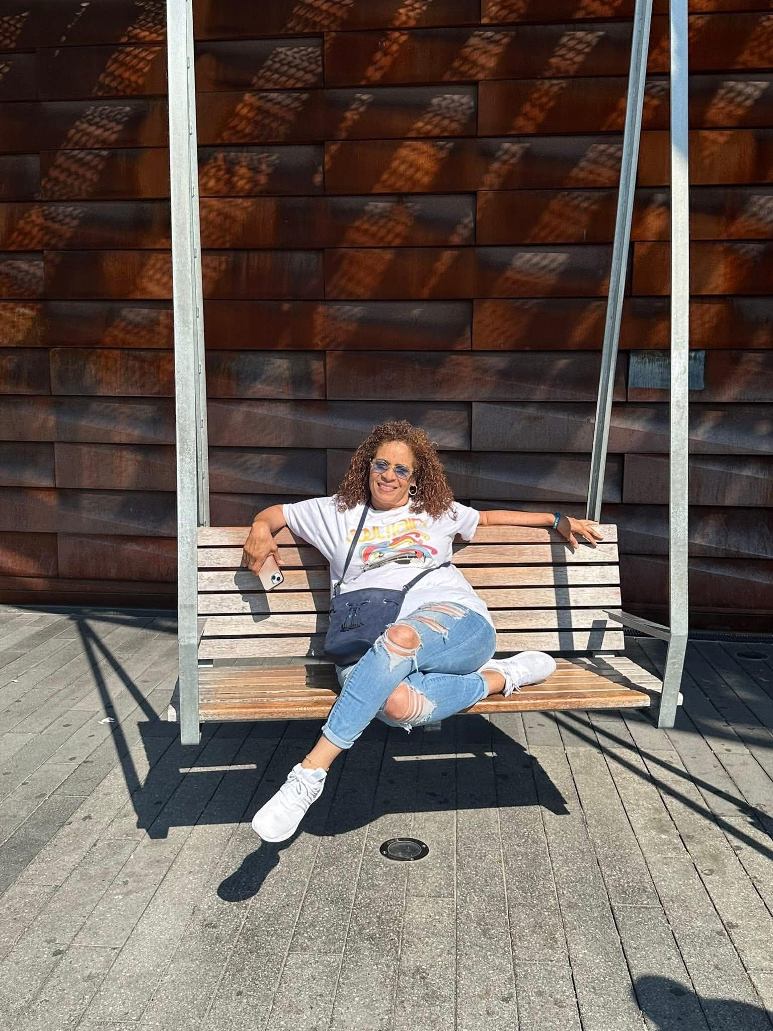 Daisy Olivo relaxing cheerfully on a suspended wooden bench swing in front of modern architecture
