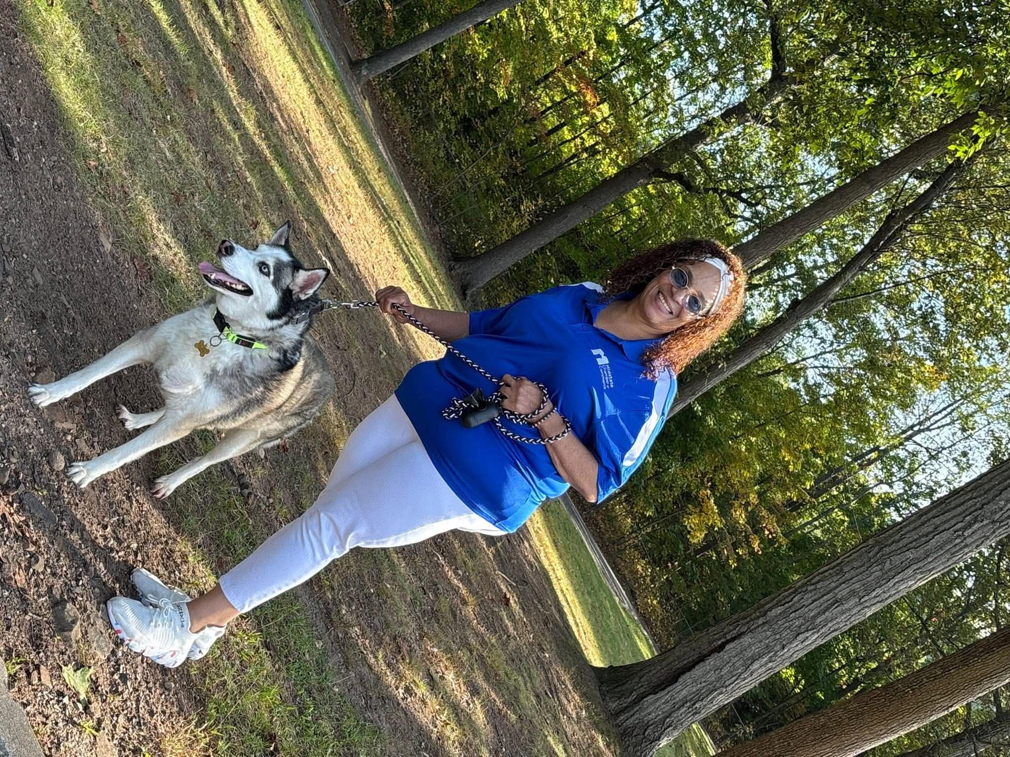 Daisy Olivo smiling outdoors in Midstate Chamber polo walking with her cheerful husky dog in a sunny wooded park