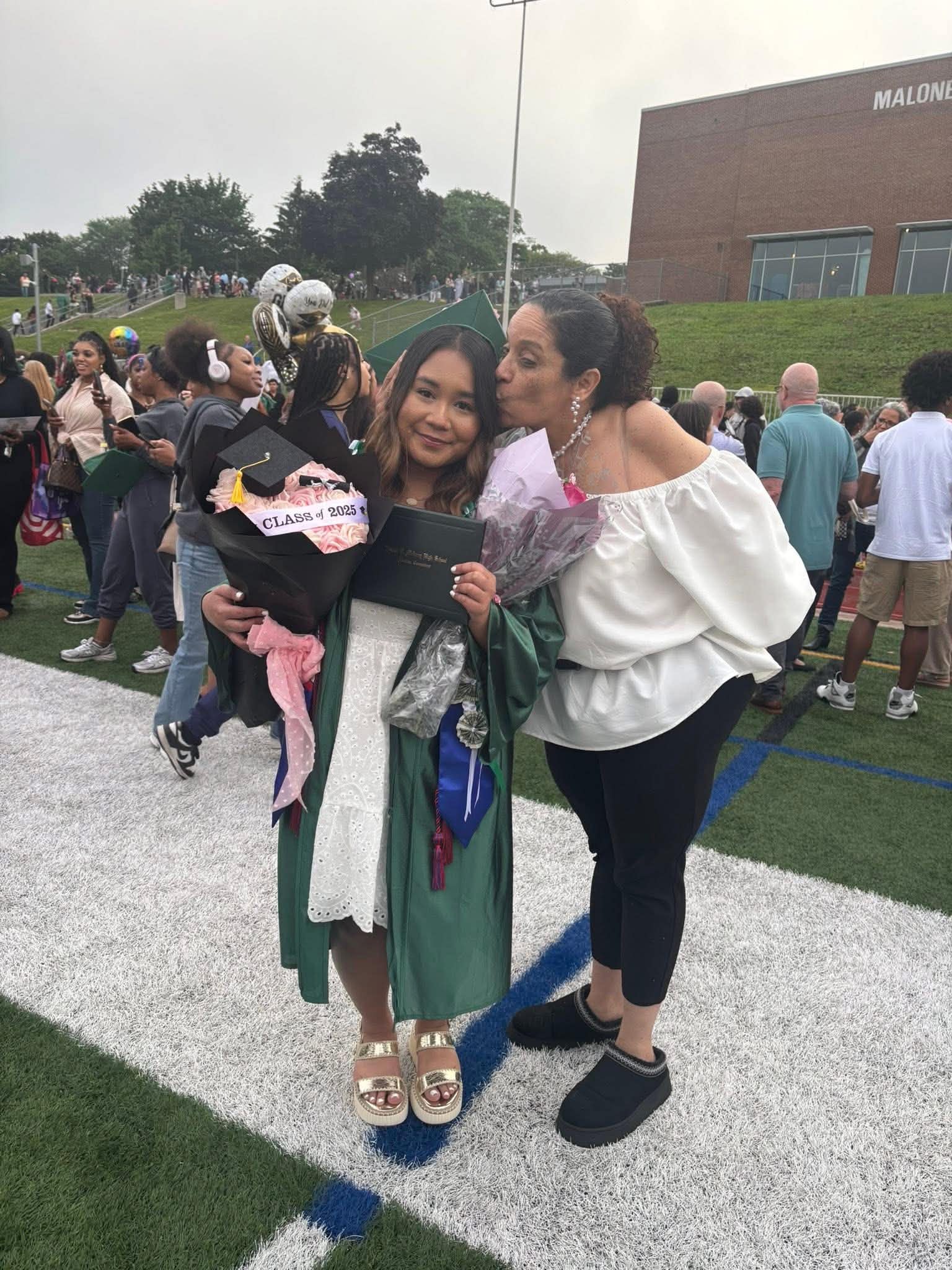 Daisy Olivo affectionately kissing a graduate holding a diploma and Class of 2025 bouquet at graduation ceremony