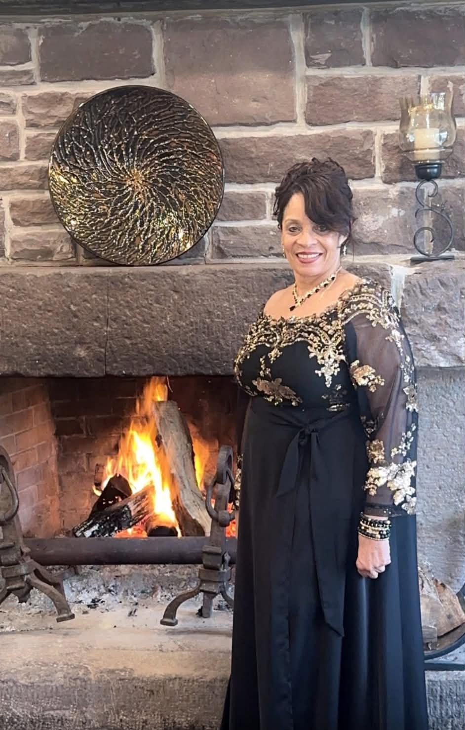 Daisy Olivo wearing an elegant black and gold gown by a stone fireplace at a community leadership gala
