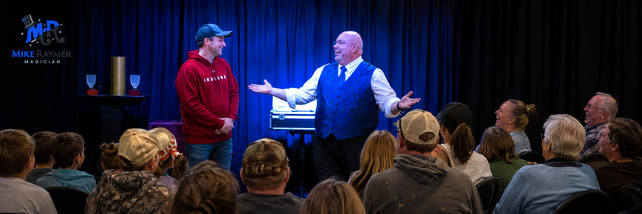 Mike Raymer performing on stage with a volunteer and audience at a family magic show