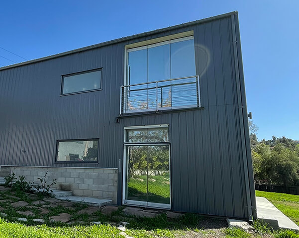 Rear view of the modern metal home with balcony, glass doors, windows, and lawn.