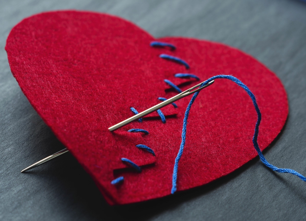 Red felt heart stitched back together with blue thread and needle, symbolising transforming past wounds into strength in the Your Sacred Scars course.