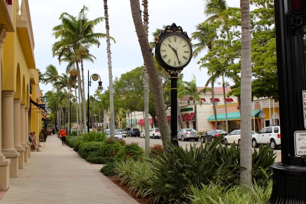 Modern daytime view of Fifth Avenue South in Naples, Florida with palm trees, storefronts, and a street clock