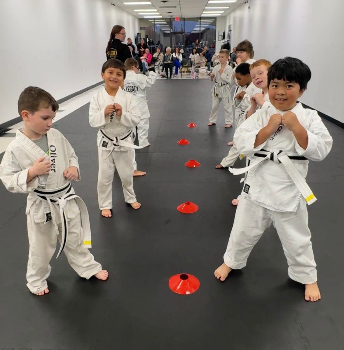 Children in white karate uniforms practicing in a dojo. Red cones mark a path.