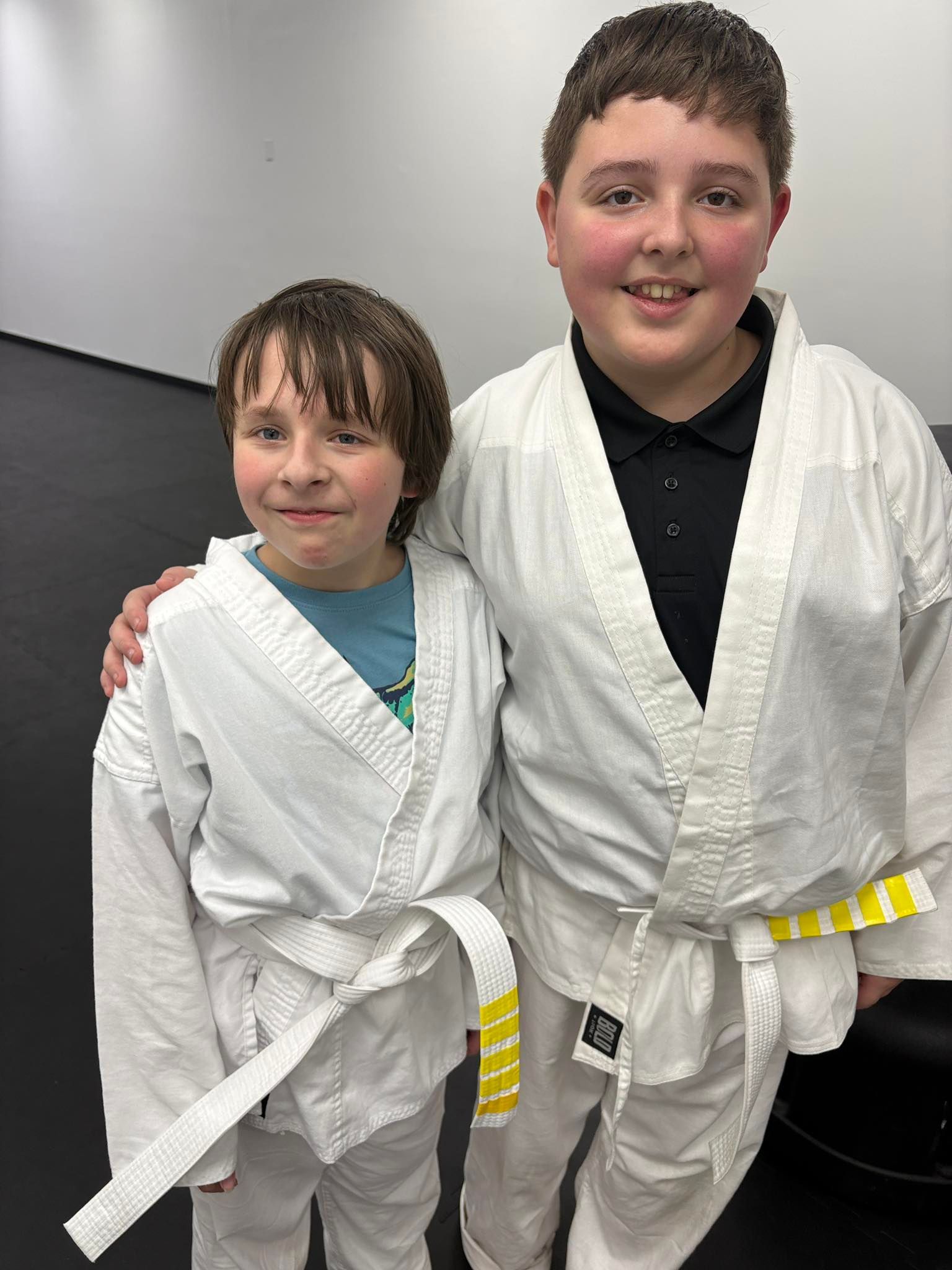 Two boys in white martial arts uniforms with yellow belts, posing indoors.