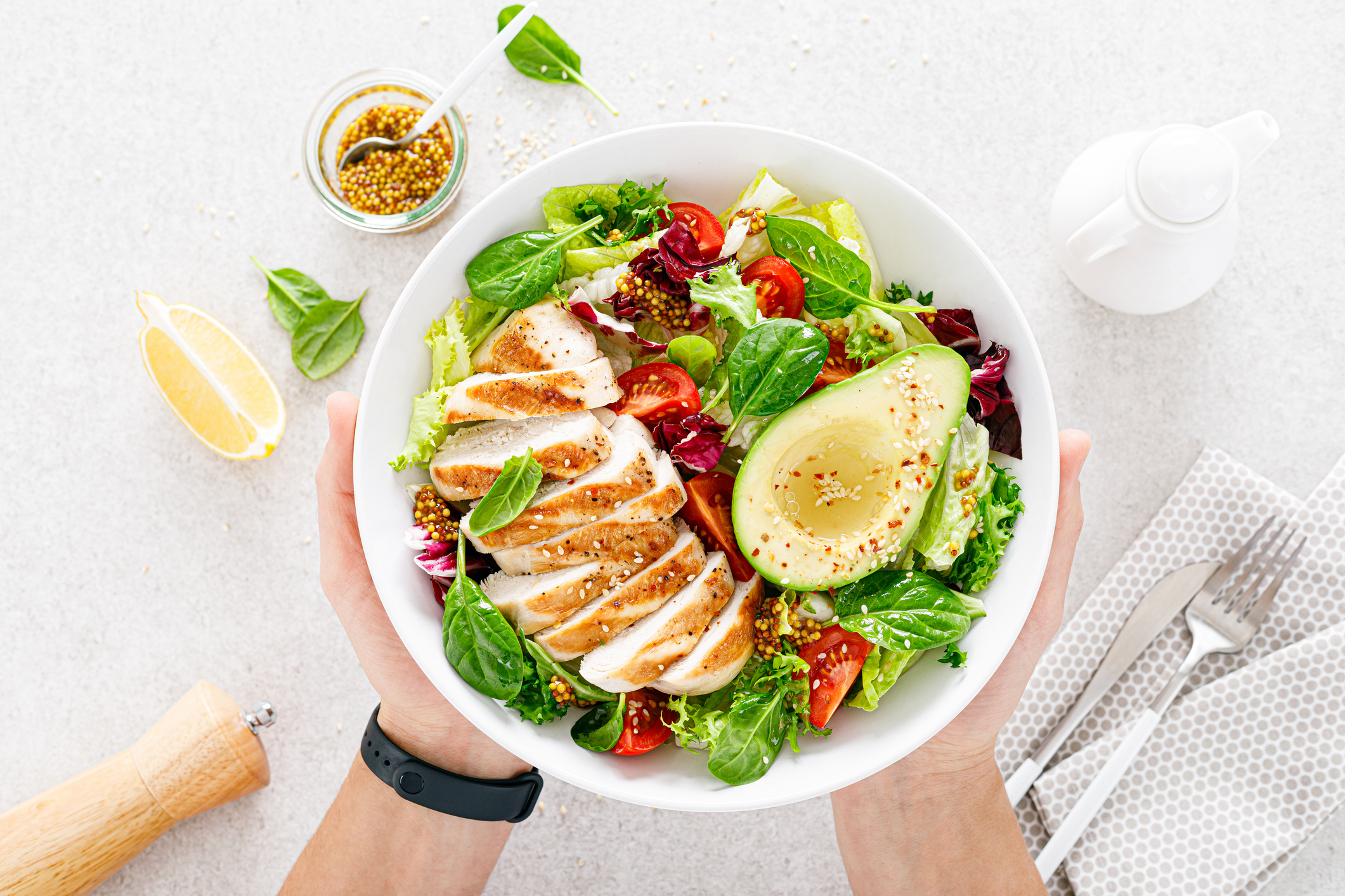 Hands holding a bowl of grilled chicken salad with avocado, tomatoes, and greens