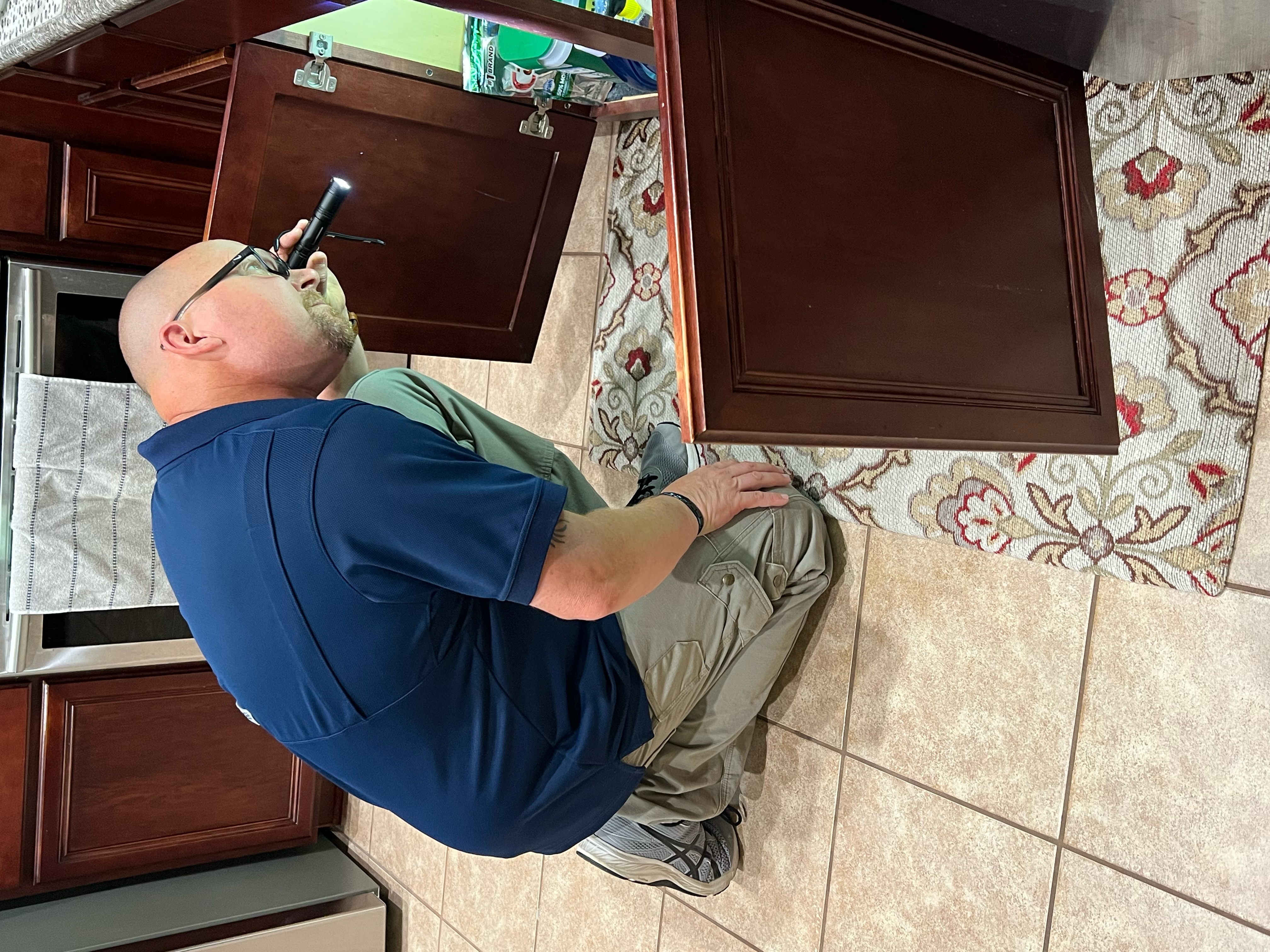 Dave Tague inspecting under-sink plumbing and cabinet with flashlight