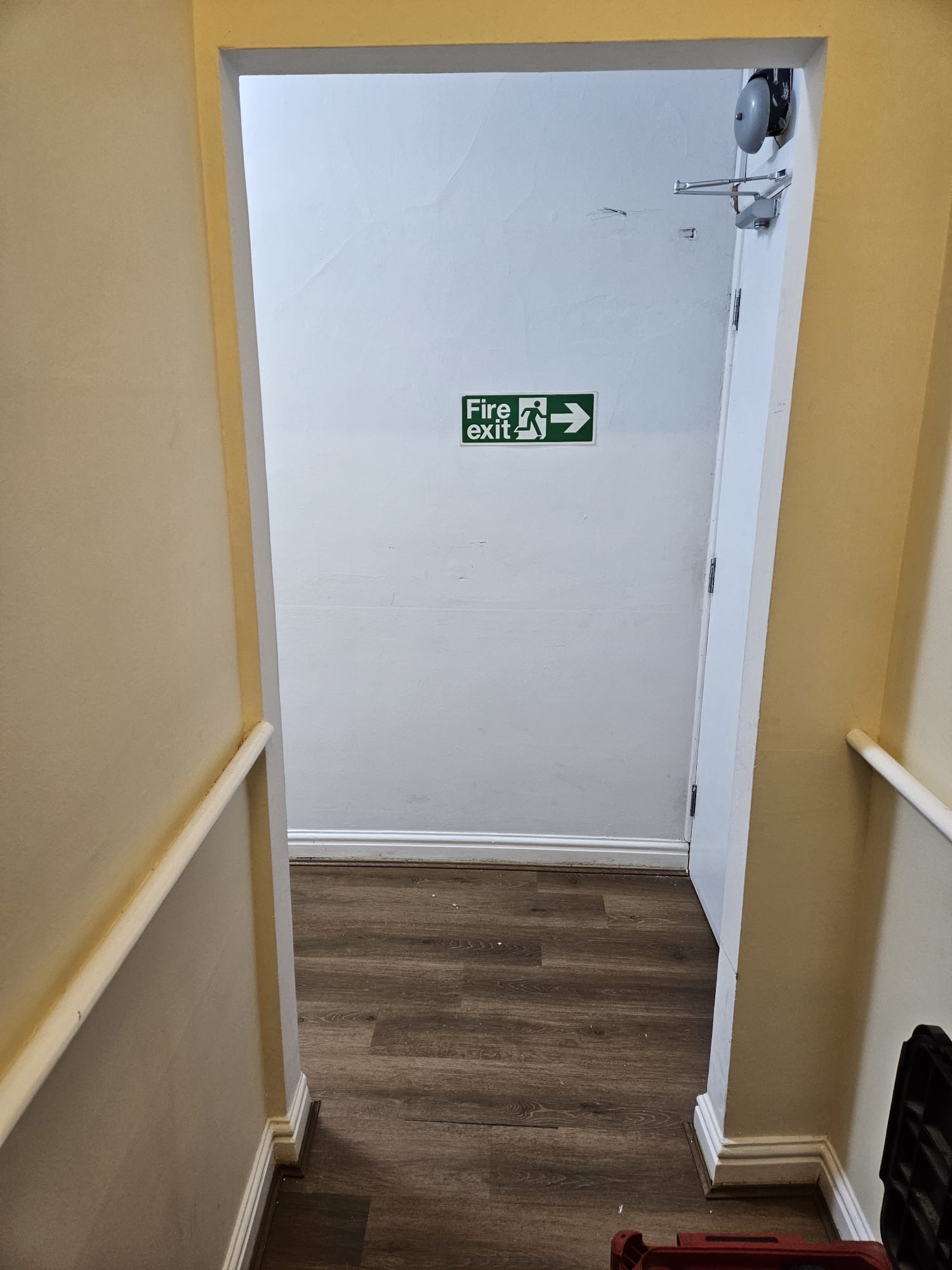 BEFORE — Plain white painted fire exit door with green Fire Exit directional signage in hallway with engineered oak flooring prior to remedial upgrade