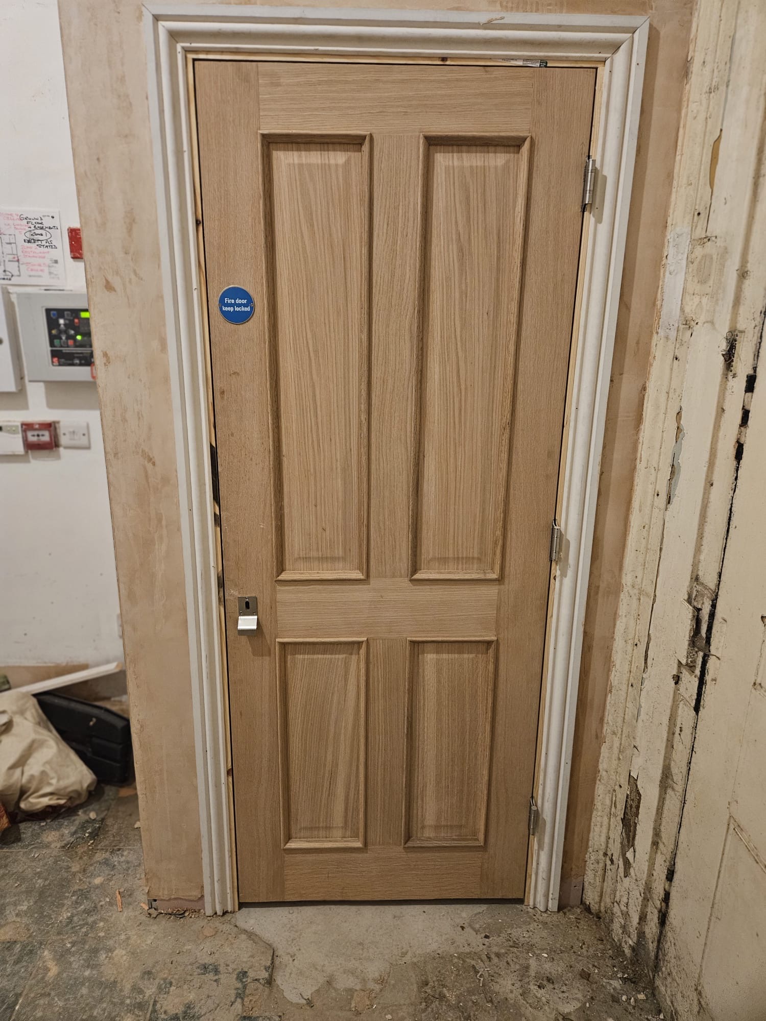 AFTER — New light oak four-panel FD30 fire door installed within primed timber frame with circular blue Fire Door Keep Shut sign and certified lever handle fitted