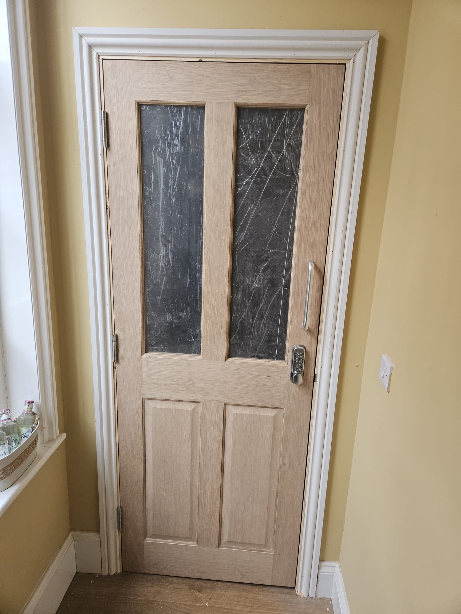 AFTER — Completed oak four-panel FD30 glazed fire door with twin vision panels, chrome D-handle and certified digital combination lock fitted within primed frame