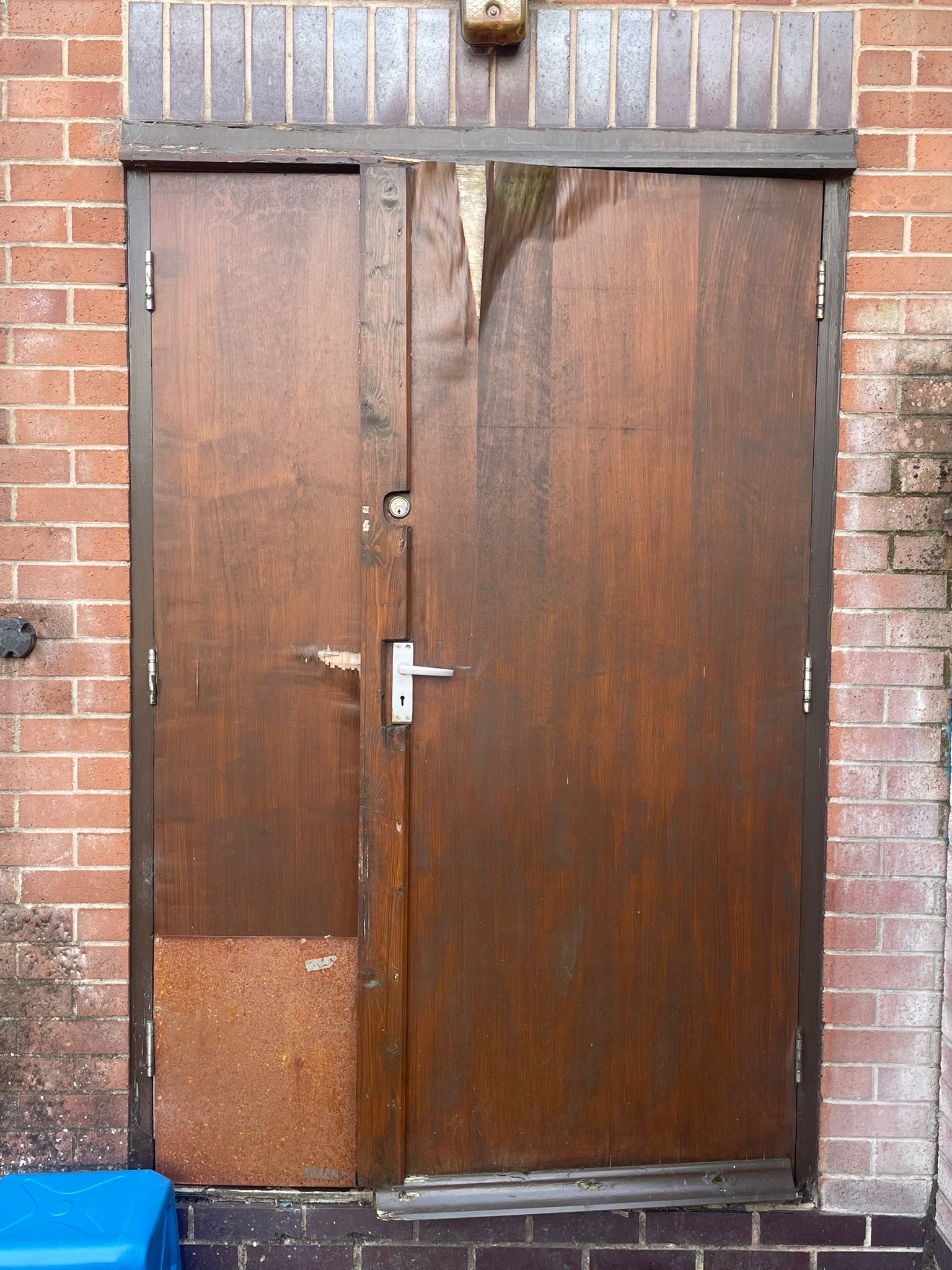 BEFORE — Weathered dark-stained double-leaf timber door set in red brick facade showing severe peeling varnish, damaged lower panel and non-compliant hardware prior to full replacement