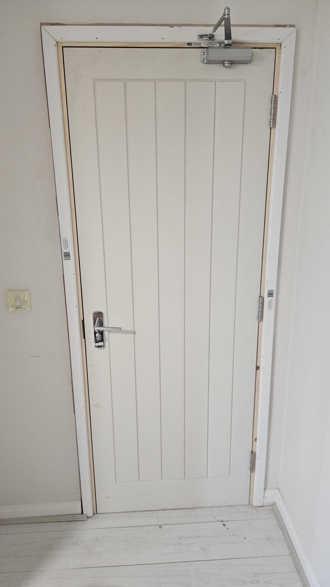 AFTER — New white vertical-panel FD30 fire door with EN 1154 overhead door closer, chrome lever handle, and new timber frame installed post-remediation