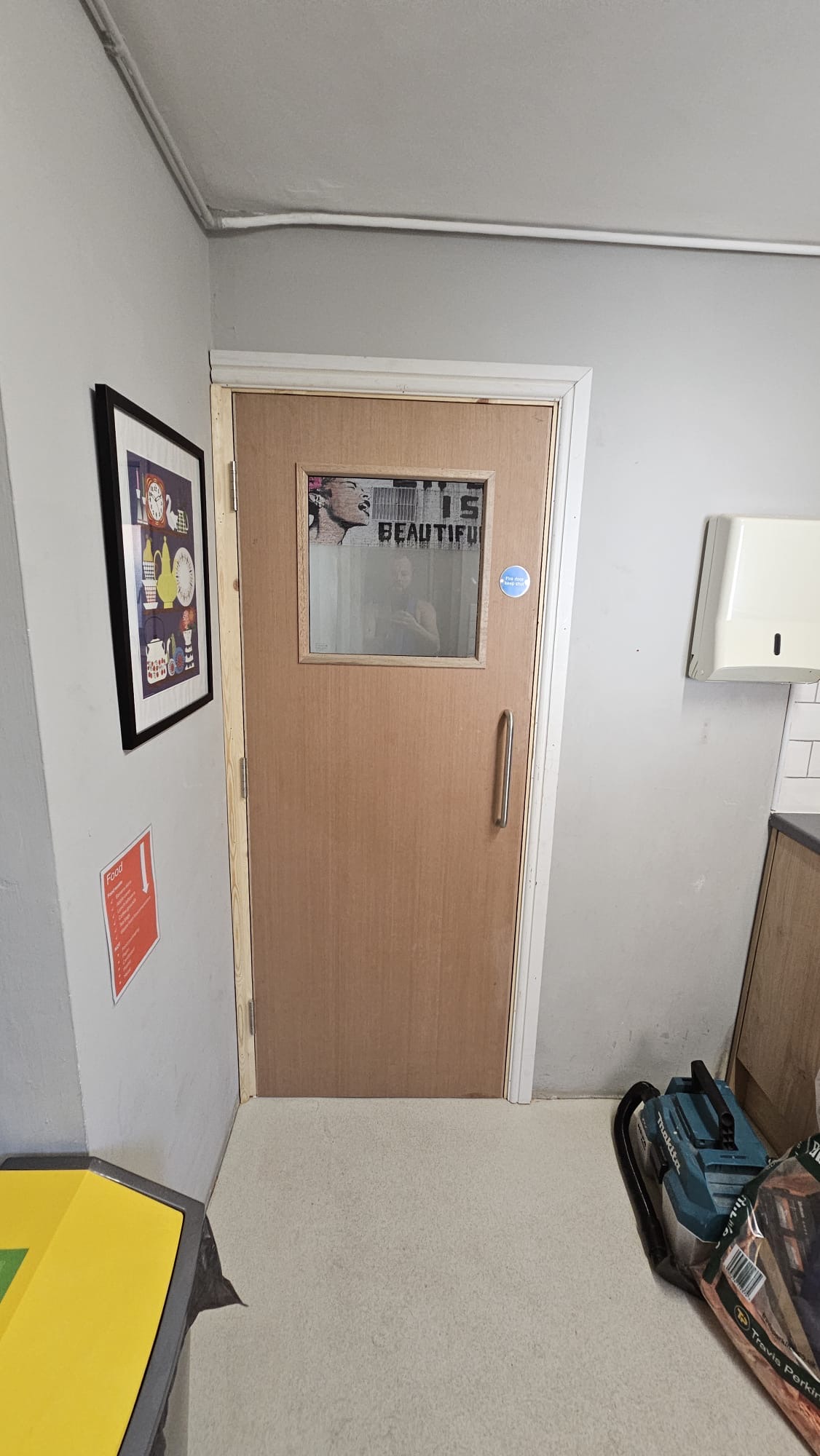 AFTER — Light oak fire door featuring rectangular vision panel, chrome D-handle, blue Fire Door Keep Shut signage installed in kitchen utility area with clean site finish