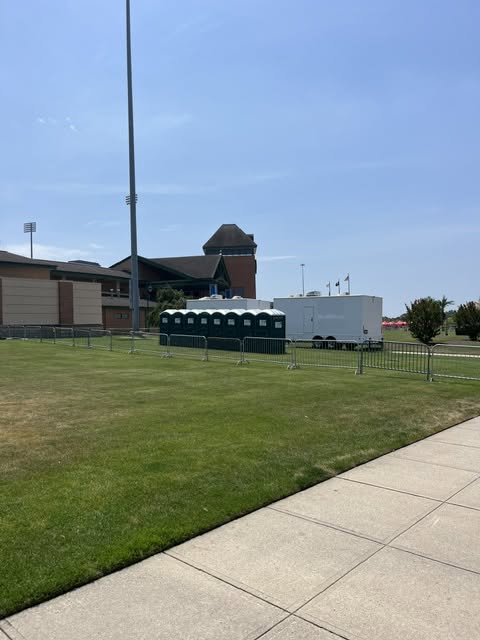 Line of green standard portable toilets and a white restroom trailer staged behind metal crowd control barricades on a field