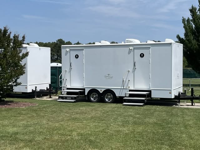 White multi-stall restroom trailer staged on lush green grass next to trees for an outdoor gathering