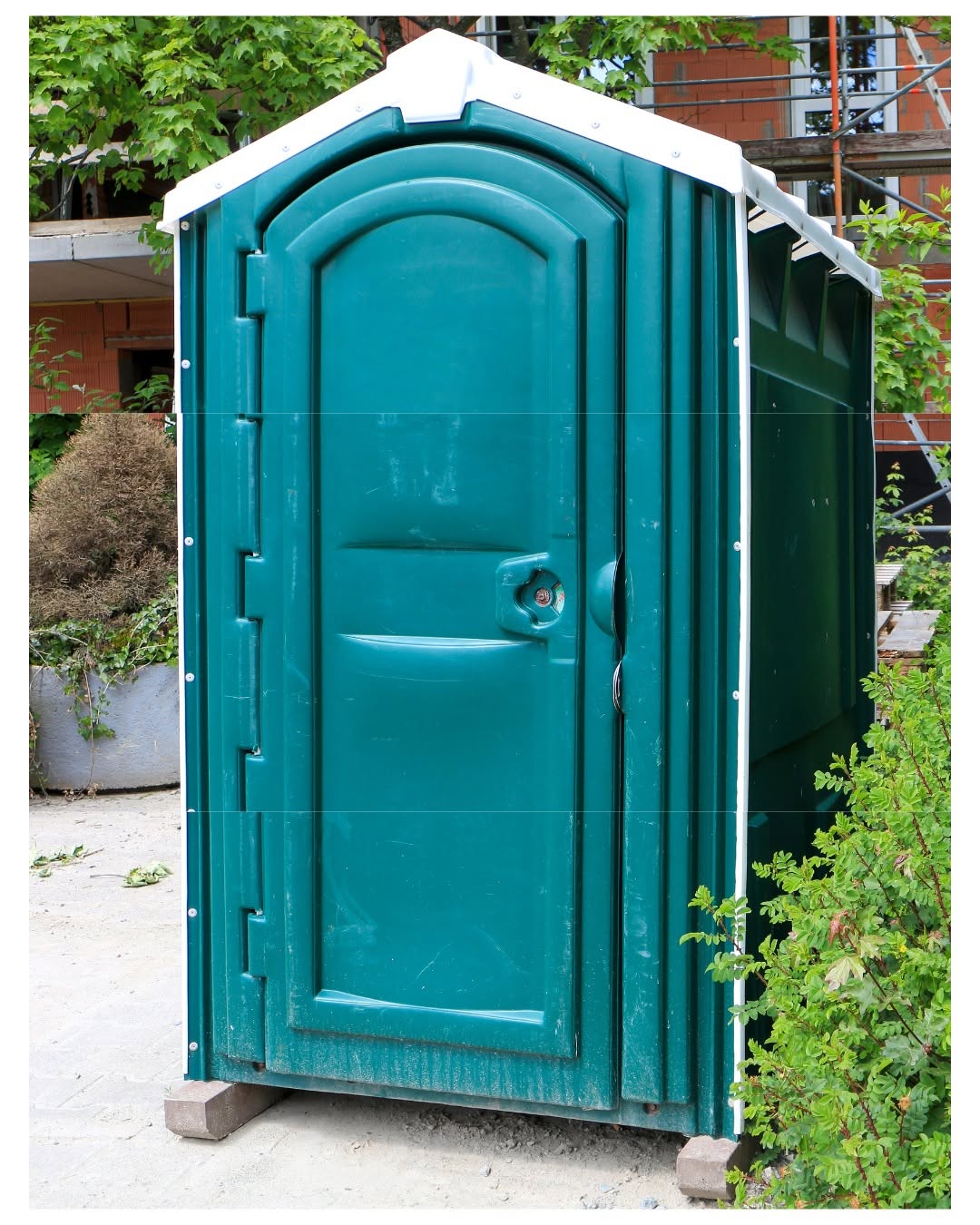 Green standard portable toilet with white roof placed on level blocks near a residential structure