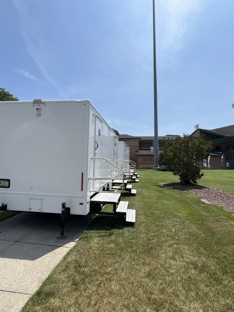 Side view of multi-stall white restroom trailers with aluminum entry steps staged along a paved walkway
